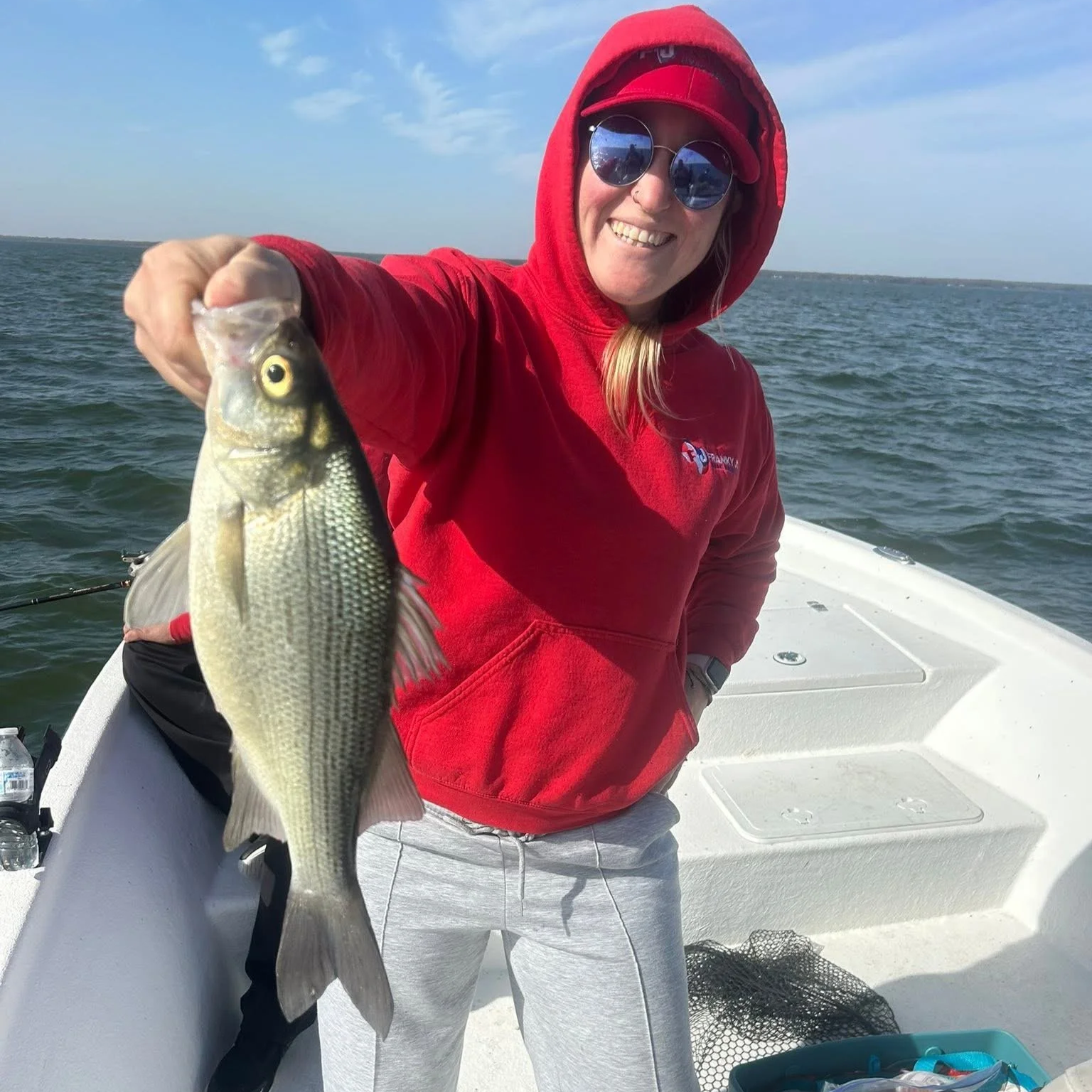 Woman in red hoodie and sunglasses holding a fish while on a boat in the water.