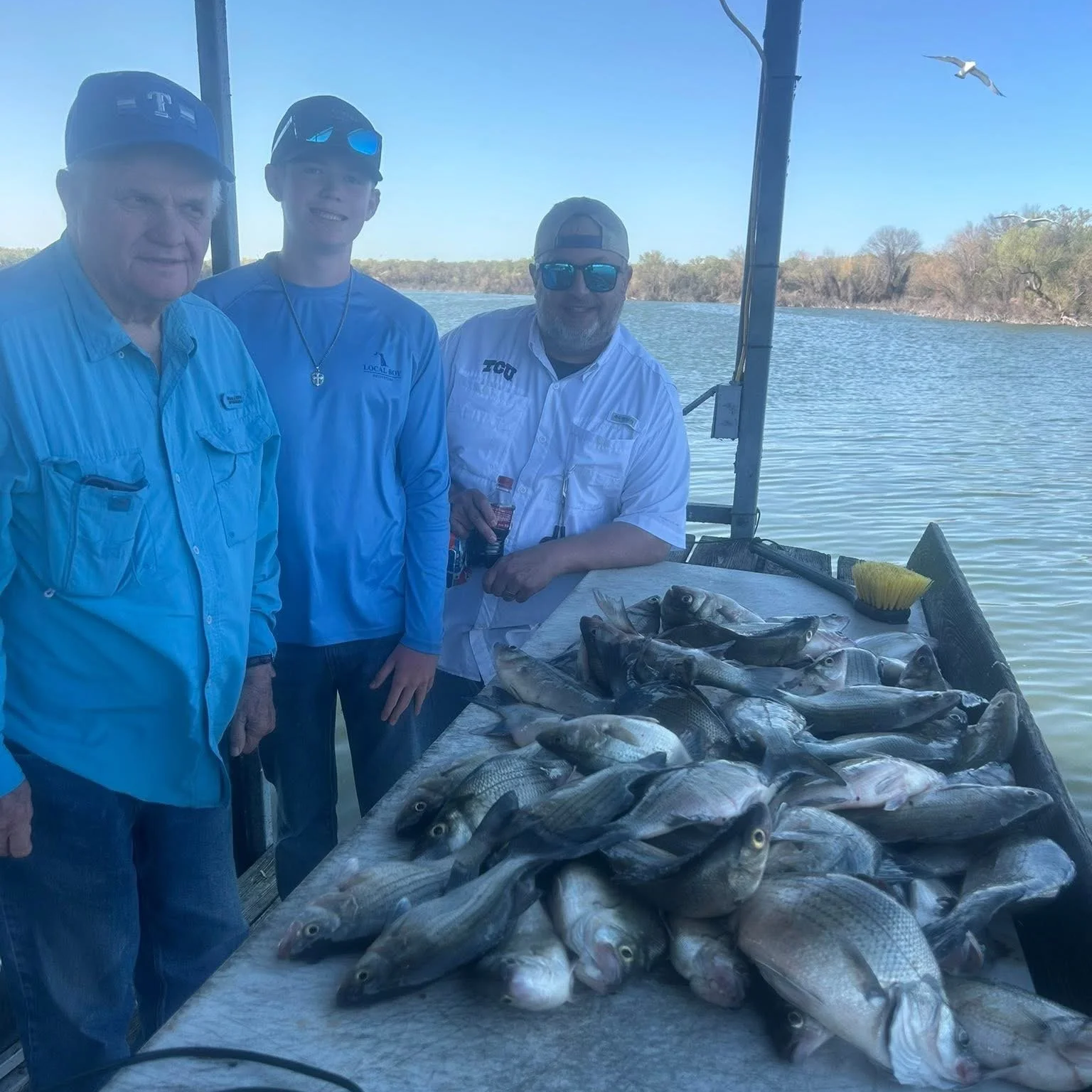 Three men standing on a boat by a table of caught fish with water and trees in the background.
