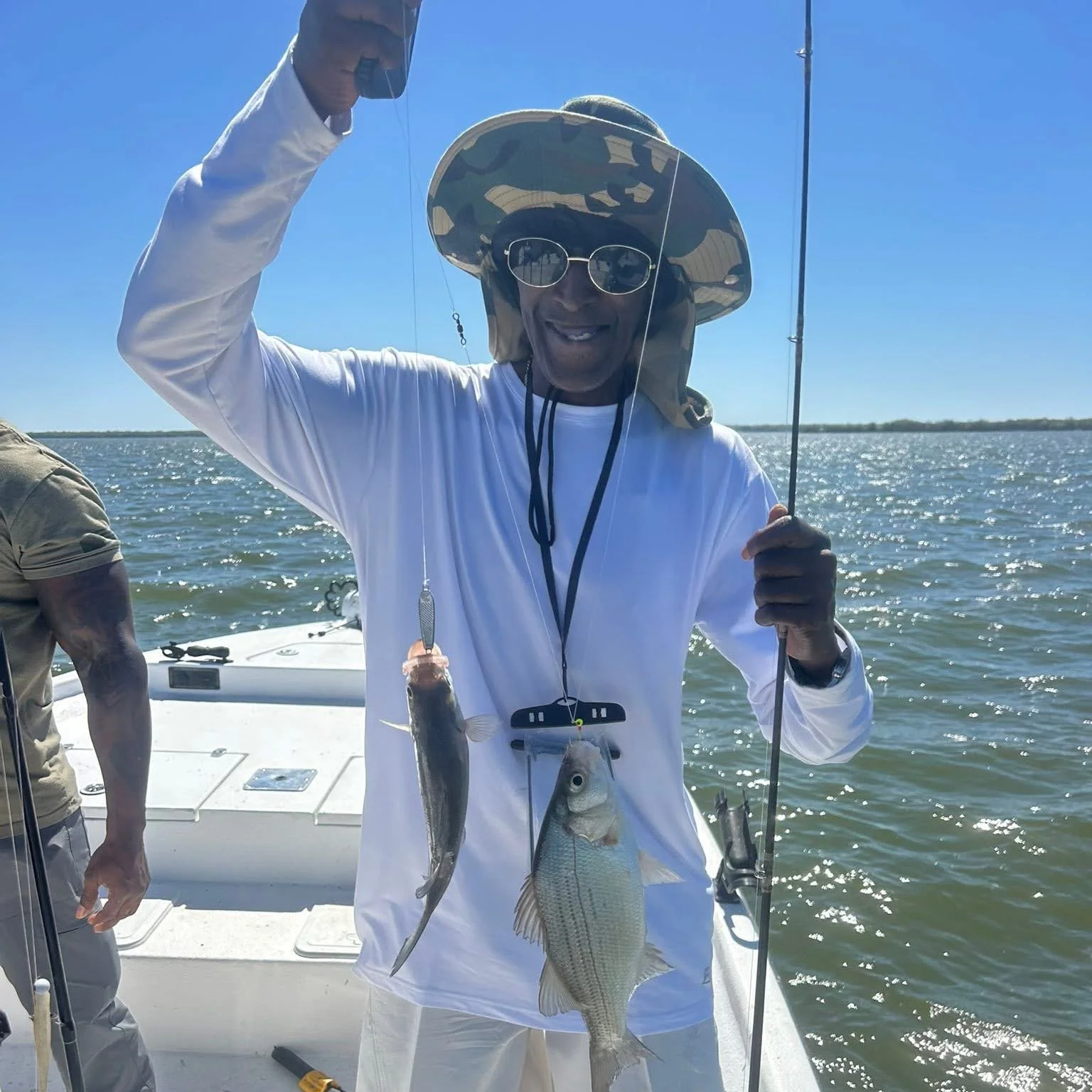 Person wearing a camo hat and sunglasses smiling while holding two fish caught during a fishing trip on a boat in a large body of water under a clear blue sky.