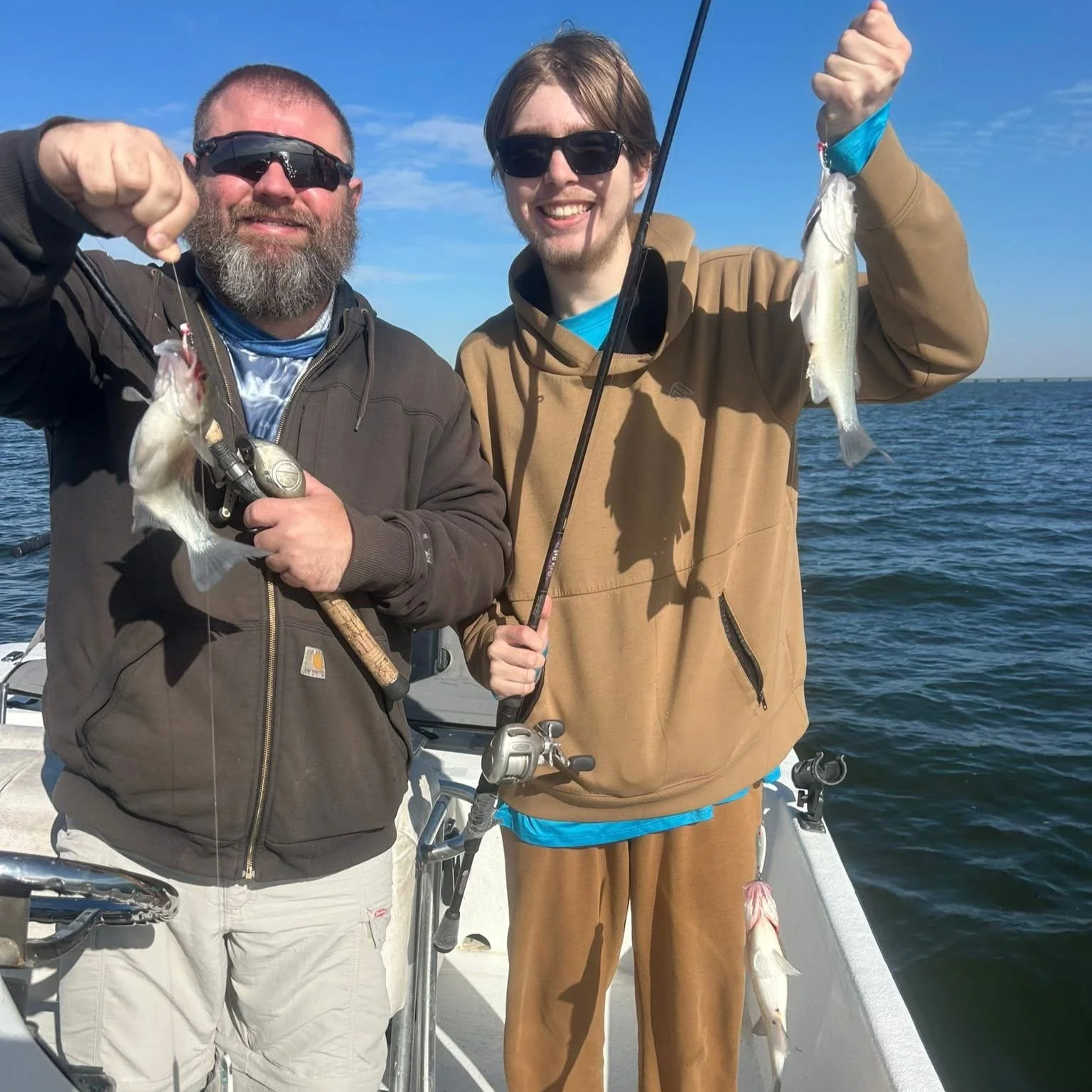 Two men on a boat holding up caught fish, smiling at the camera with water and blue sky in the background.