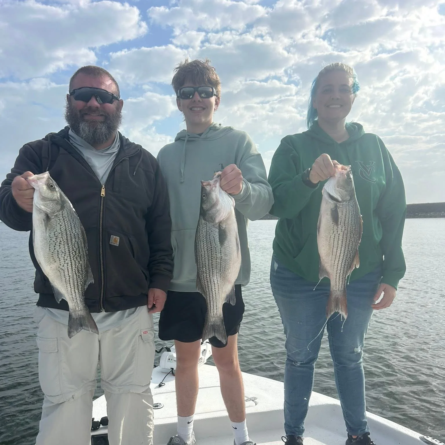 Three people standing on a boat holding large fish they caught, with water and a cloudy sky in the background.