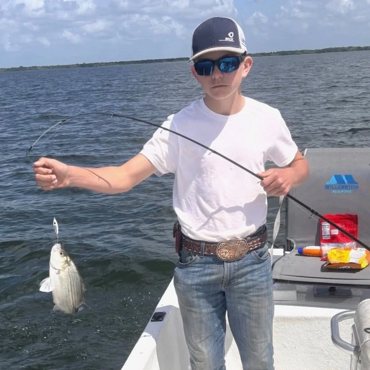 Young boy wearing sunglasses and a baseball cap holding a fishing rod and displaying a caught fish on a boat in a lake.