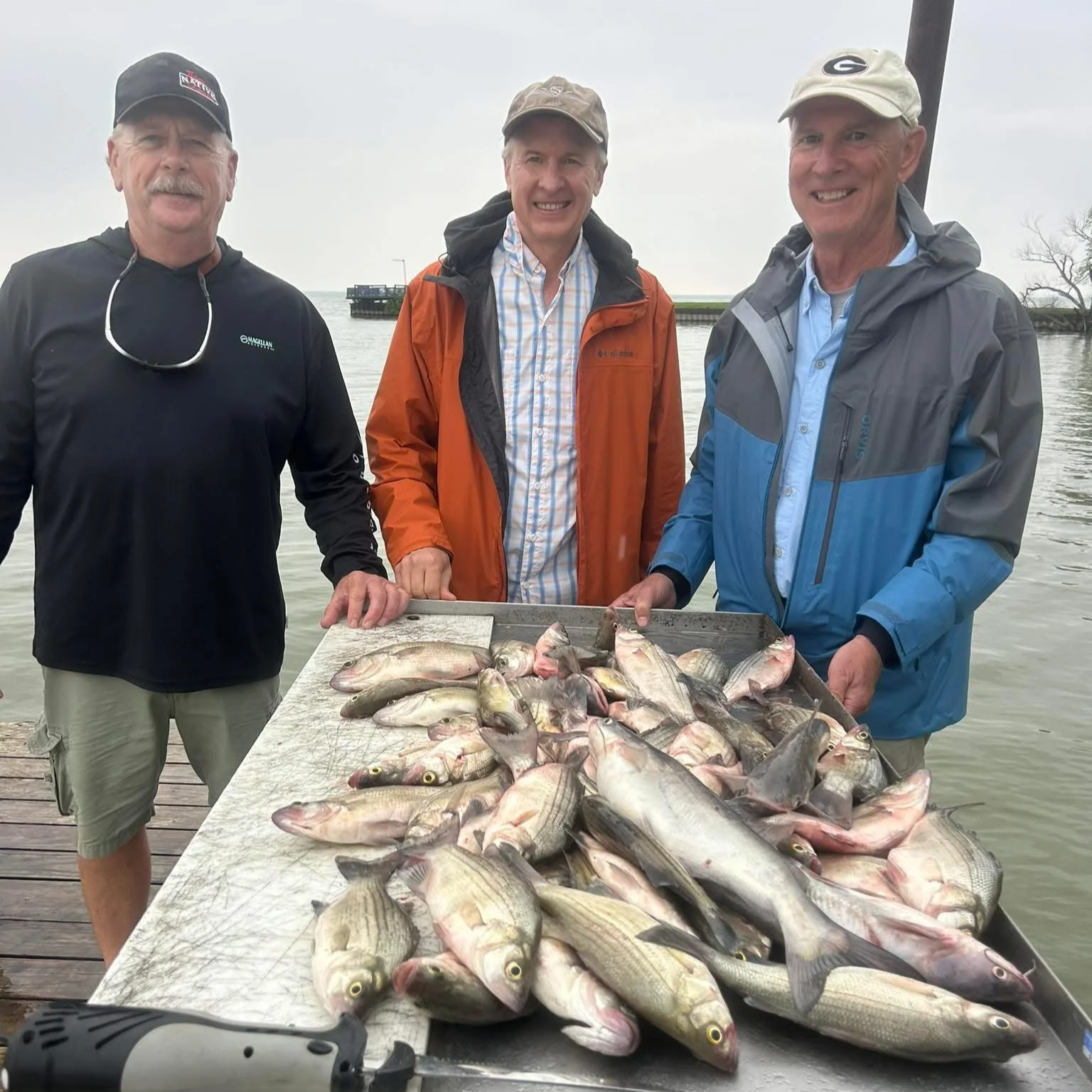 Three men standing behind a table full of freshly caught fish, smiling outdoors by a body of water on a cloudy day.