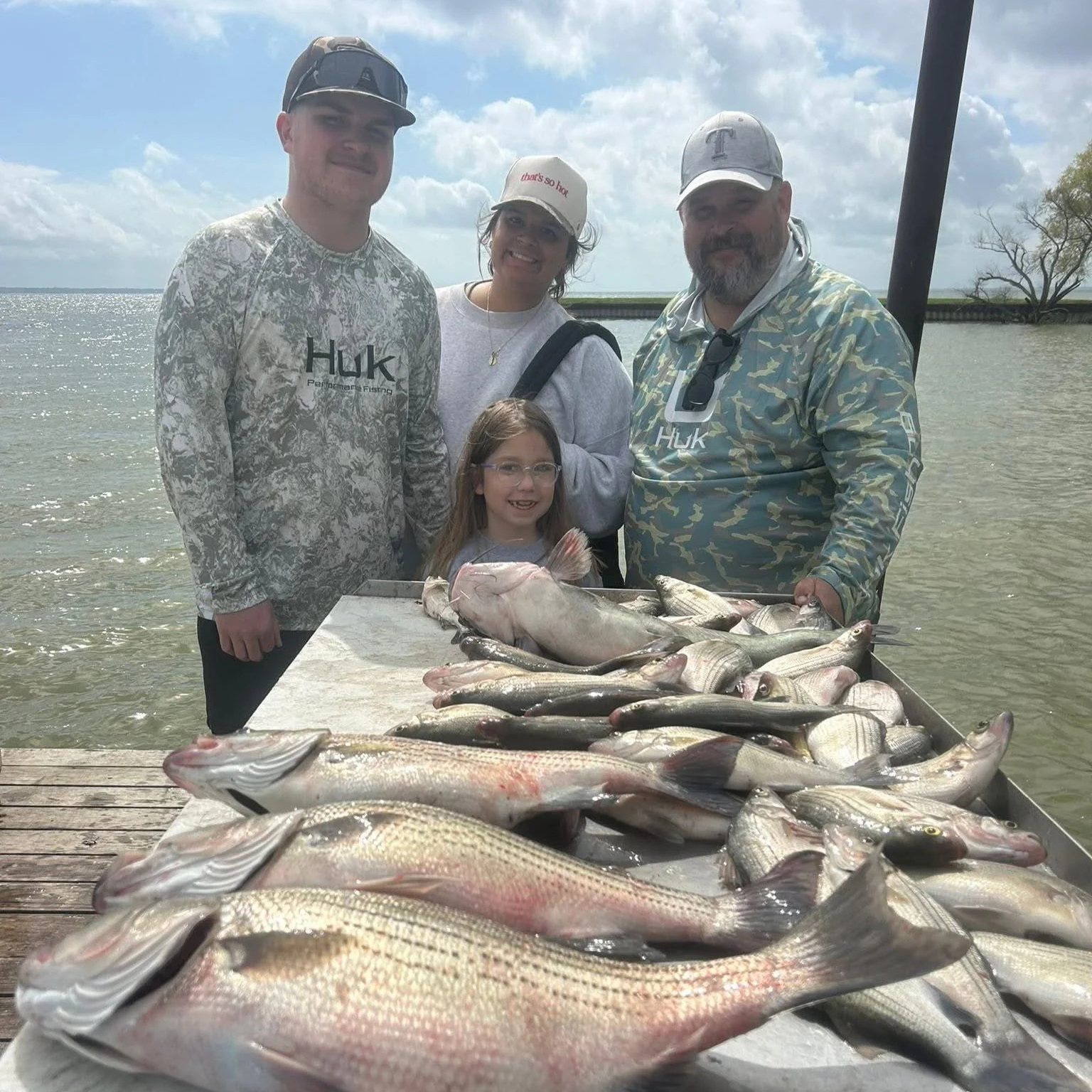 A group of five people, including two children, standing behind a table filled with fish, outdoors near a body of water on a partly cloudy day.