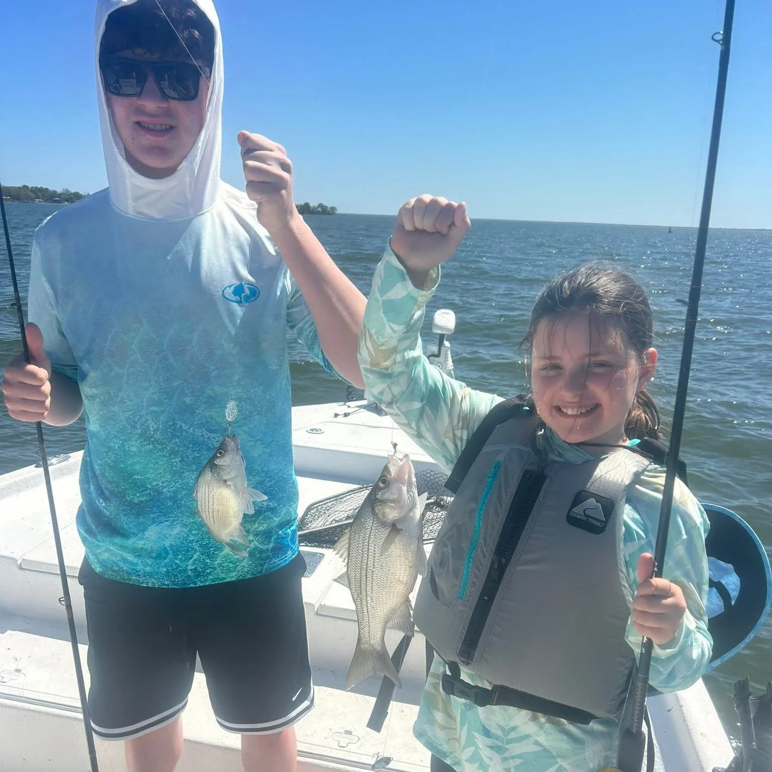 Two children on a boat holding up caught fish, with one child wearing sunglasses and a hooded shirt, the other smiling and holding a fishing rod, with water and a clear sky in the background.