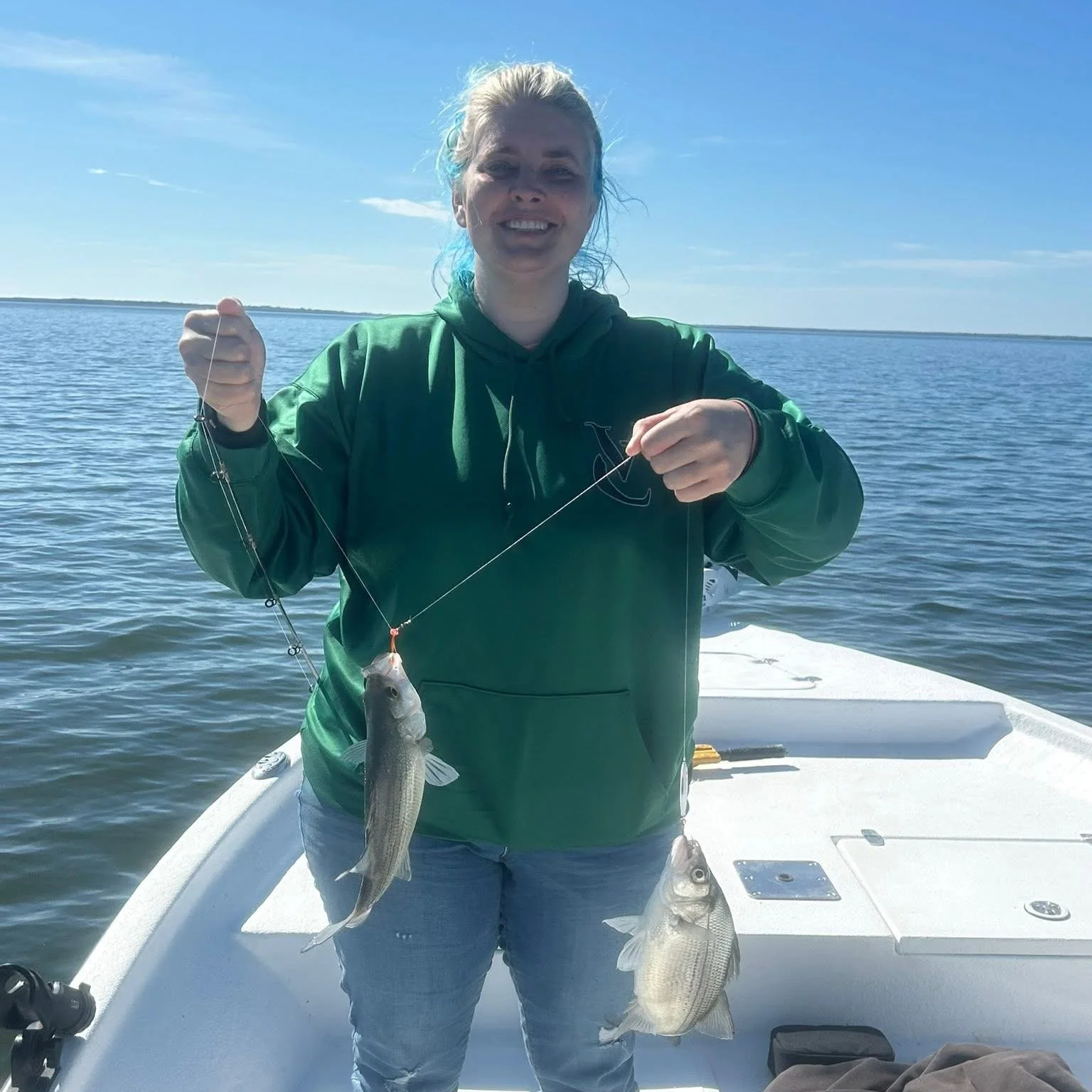 A woman on a boat holding two fish she caught while fishing under a clear blue sky.