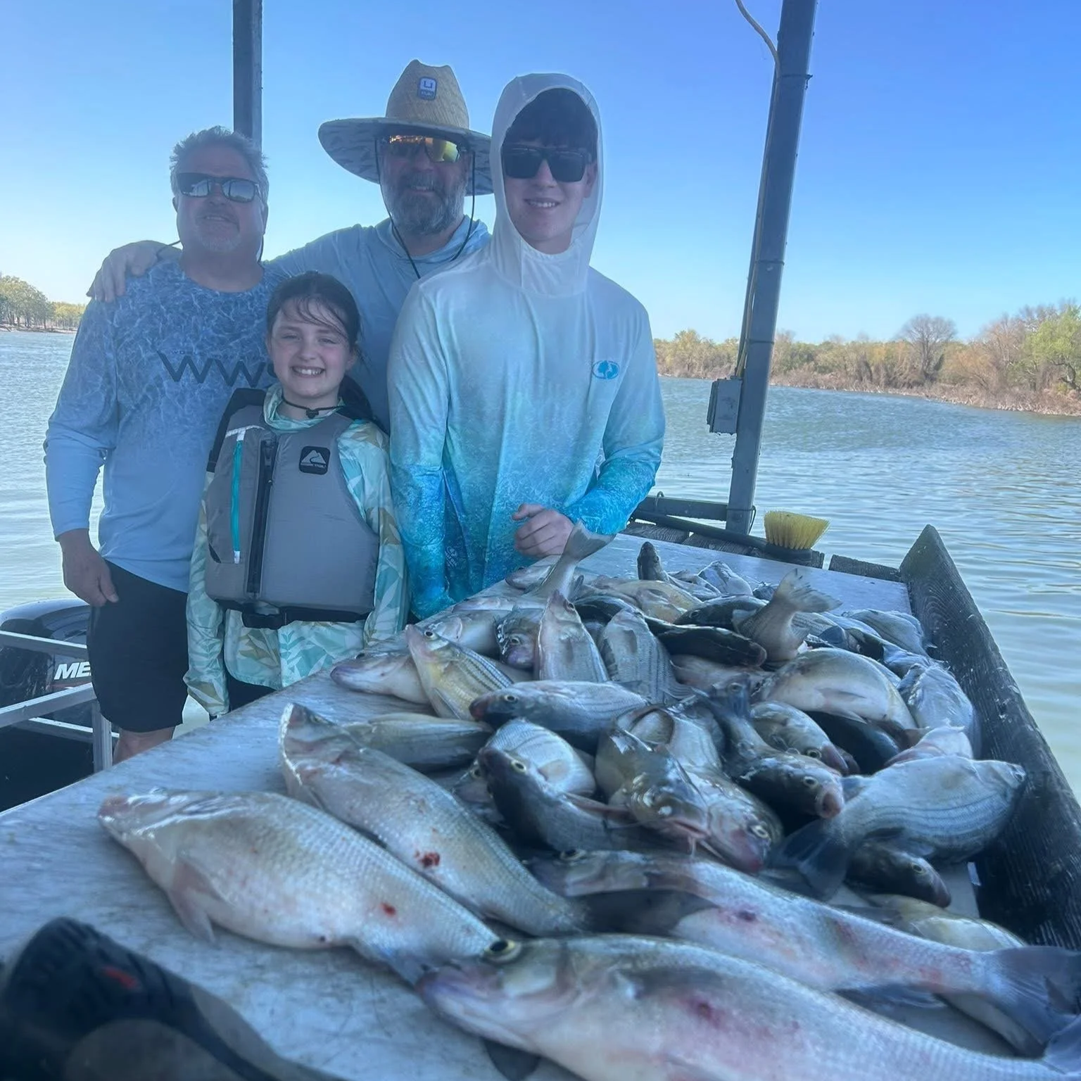 Family of four on a boat near a lake, posing behind a large pile of caught fish during daytime.