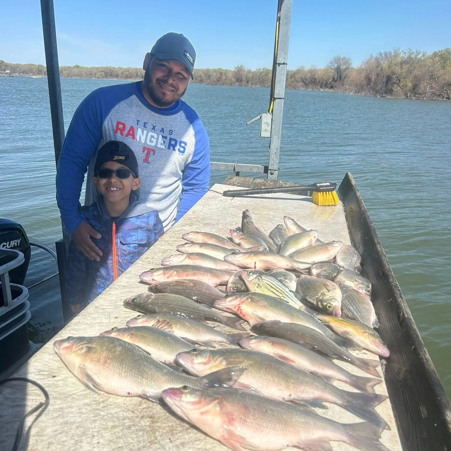 A man and a boy on a boat with a fishing rod, smiling next to a table full of various caught fish, with a body of water and trees in the background.