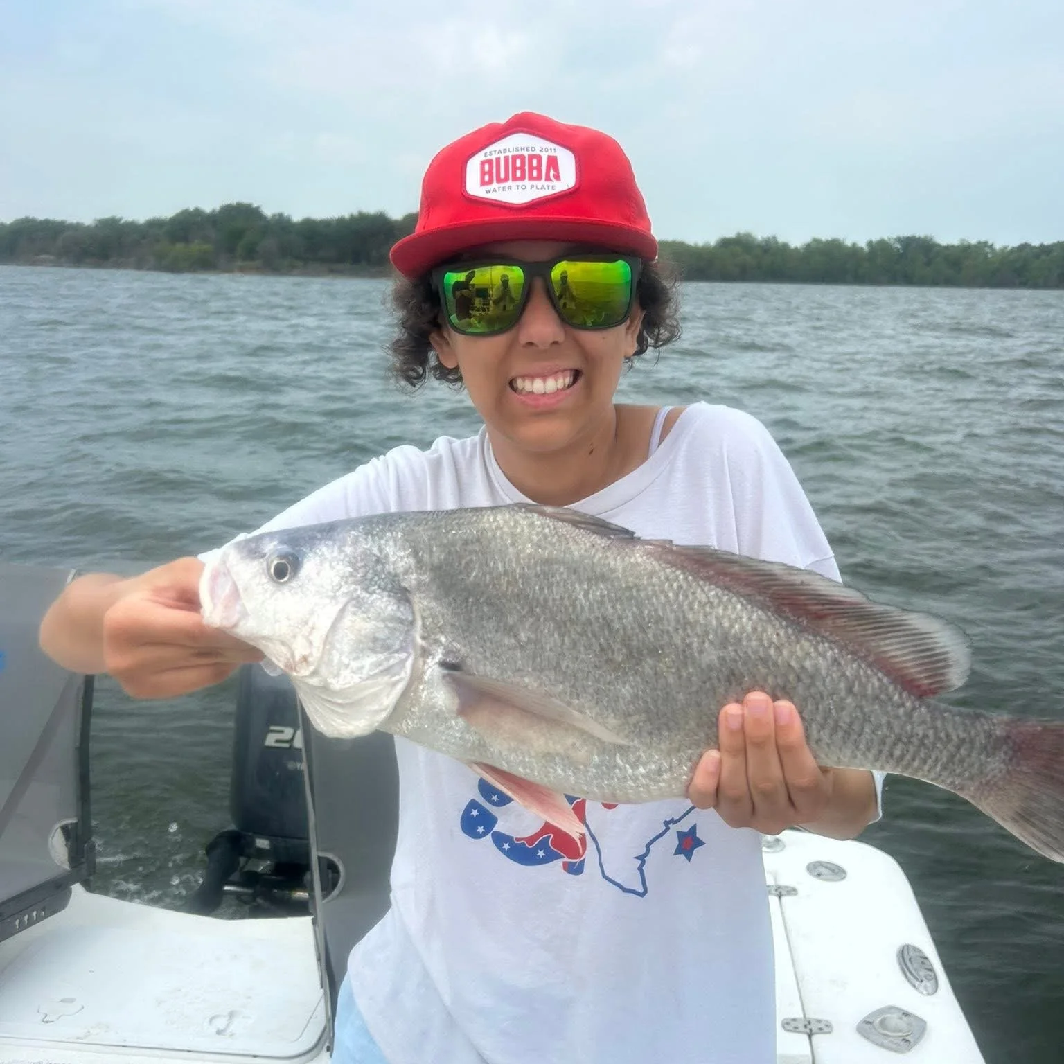 A person wearing sunglasses and a red cap holding a large fish on a boat in a body of water with trees in the background.