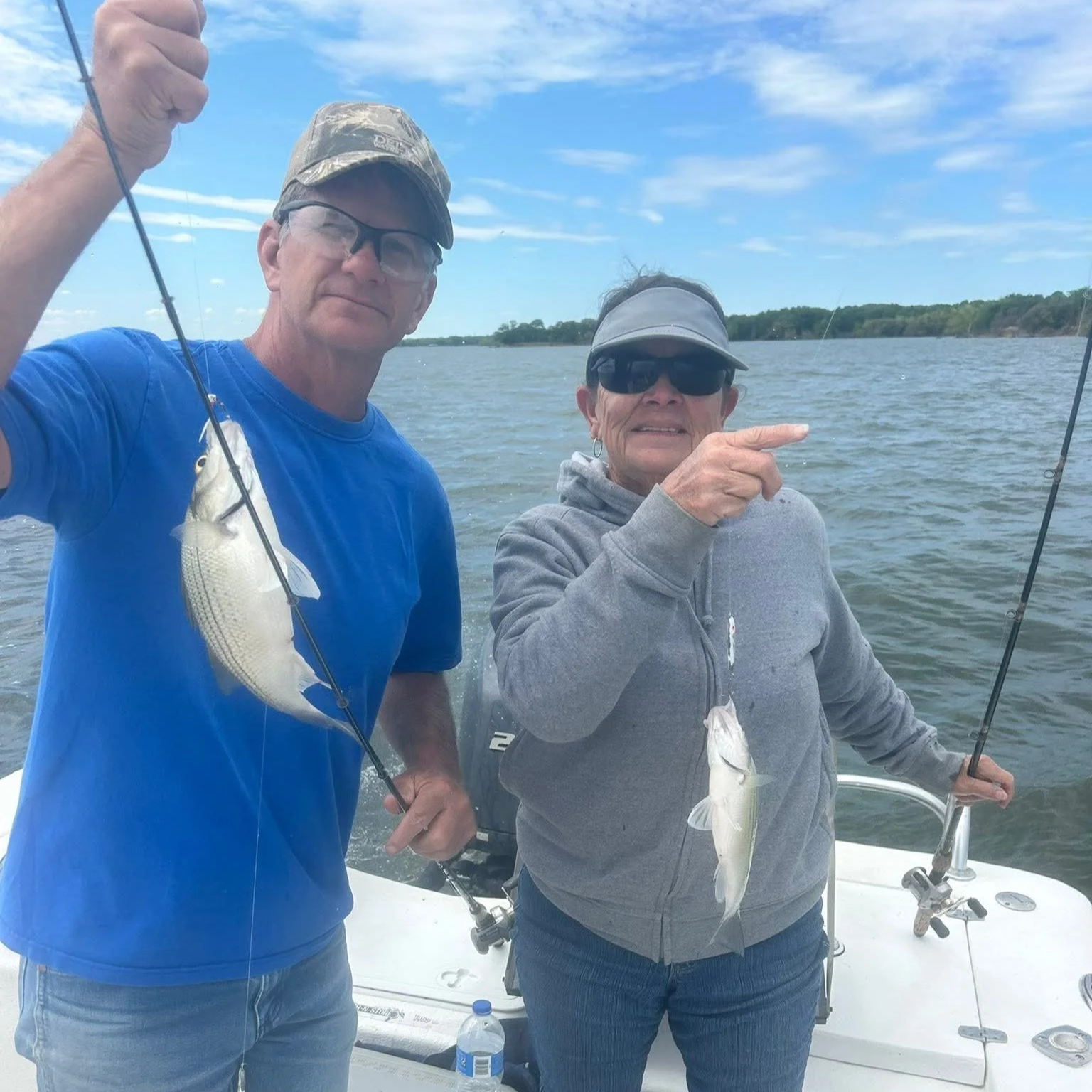 Two people on a boat holding fishing rods, showing their caught fish, with water and a partly cloudy sky in the background.