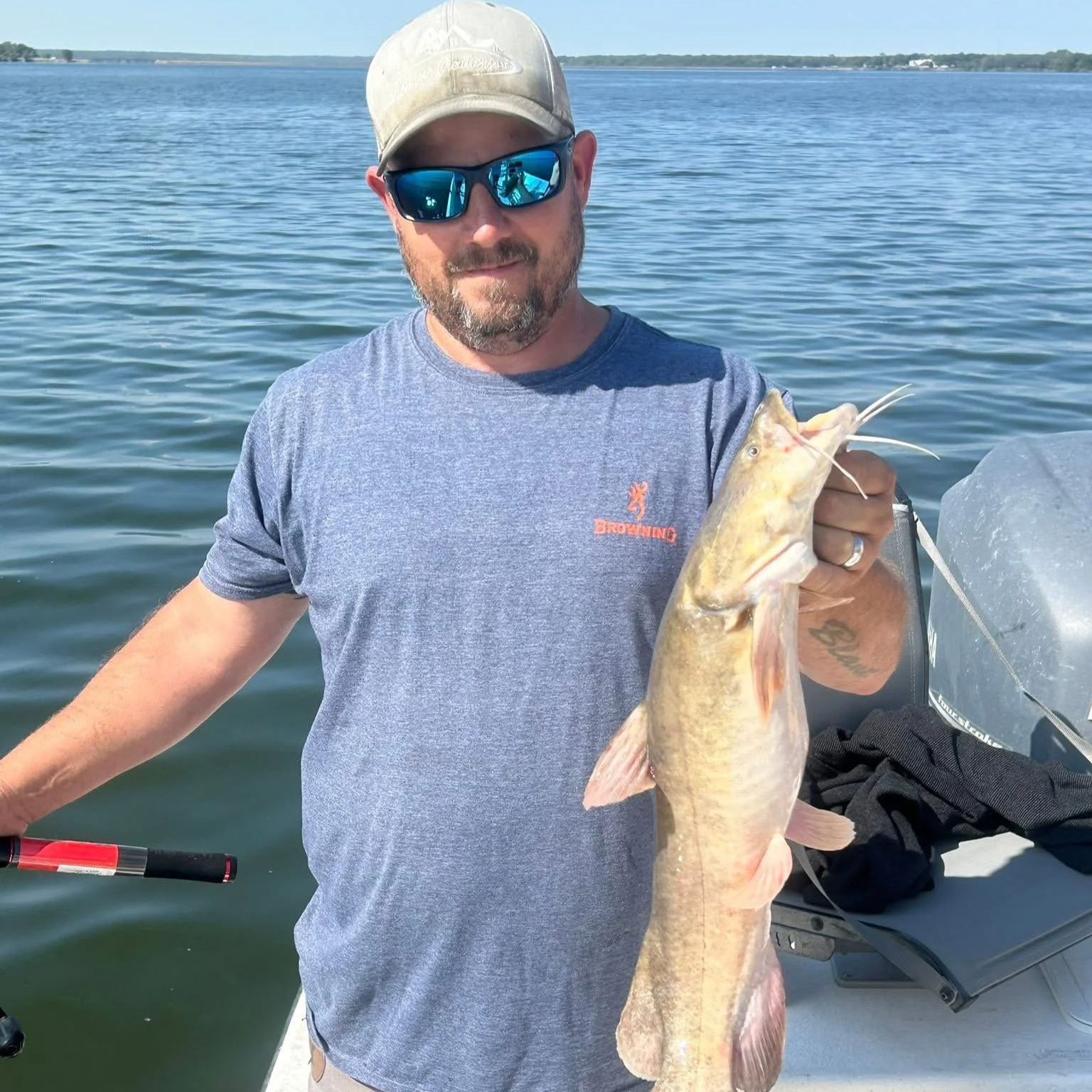 A man with a beard wearing sunglasses, a cap, and a gray t-shirt holding a large fish on a boat in a body of water.