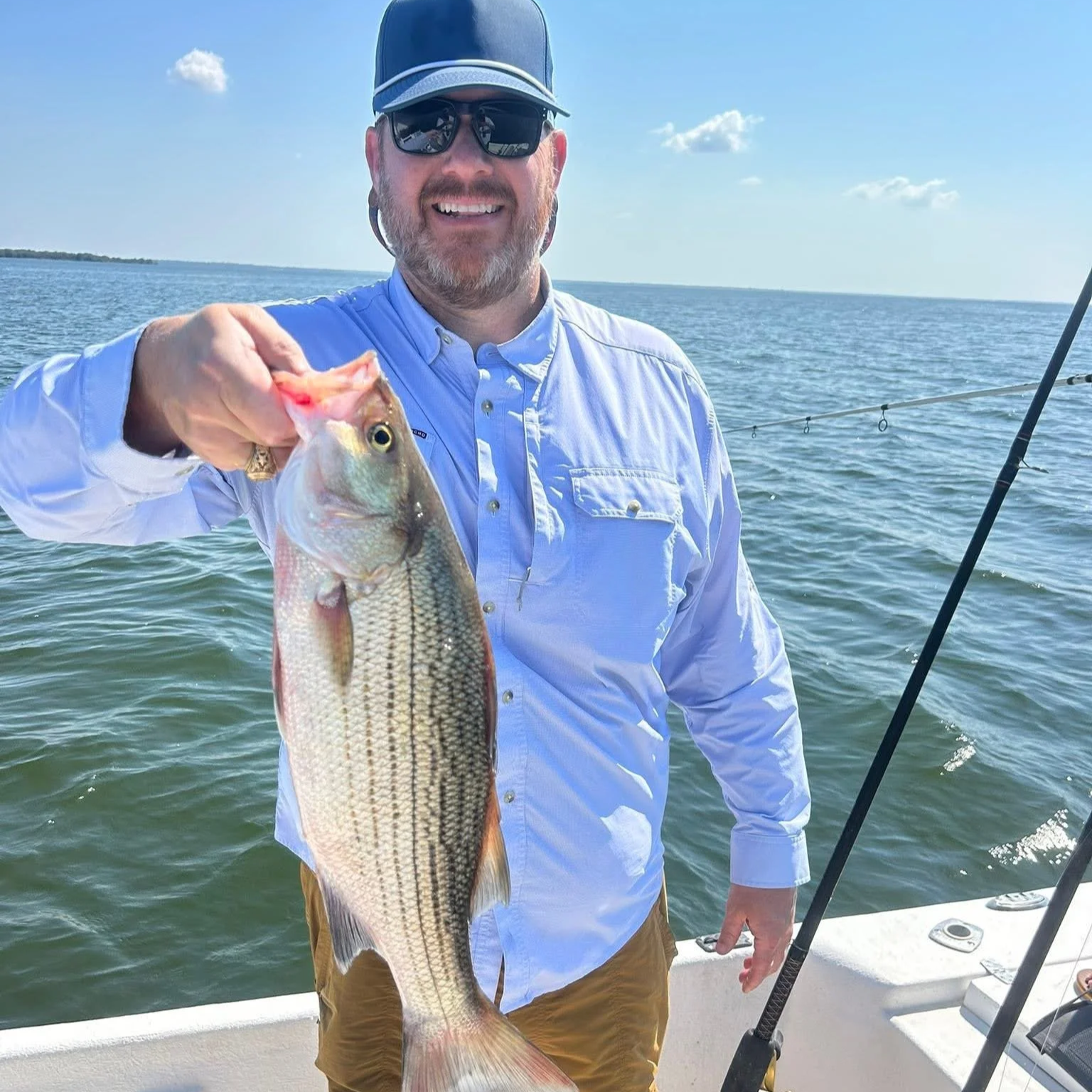 A man with sunglasses and a blue cap holding a largemouth bass on a boat in a large body of water, smiling at the camera, with fishing gear and a fishing rod visible.