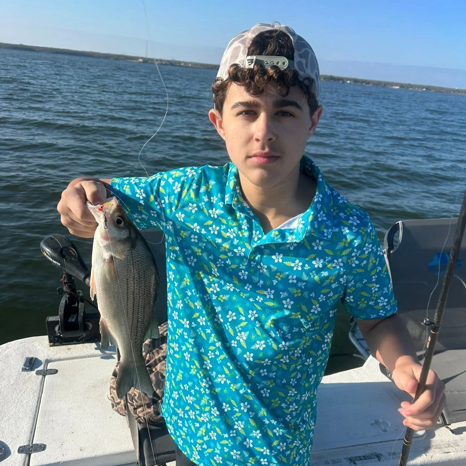 A young man on a boat holding a freshly caught fish, with water and a distant shoreline in the background.