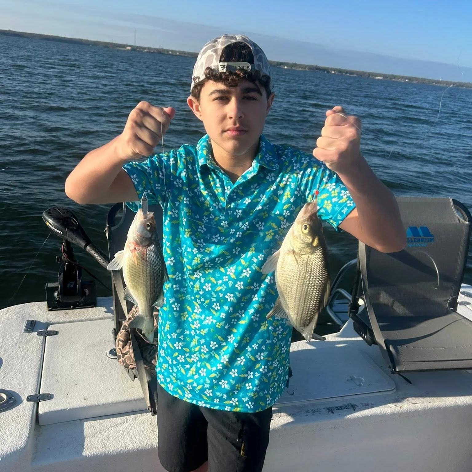 A boy on a boat holding two fishing rods with fish caught on the lines, with water and a distant shoreline in the background.