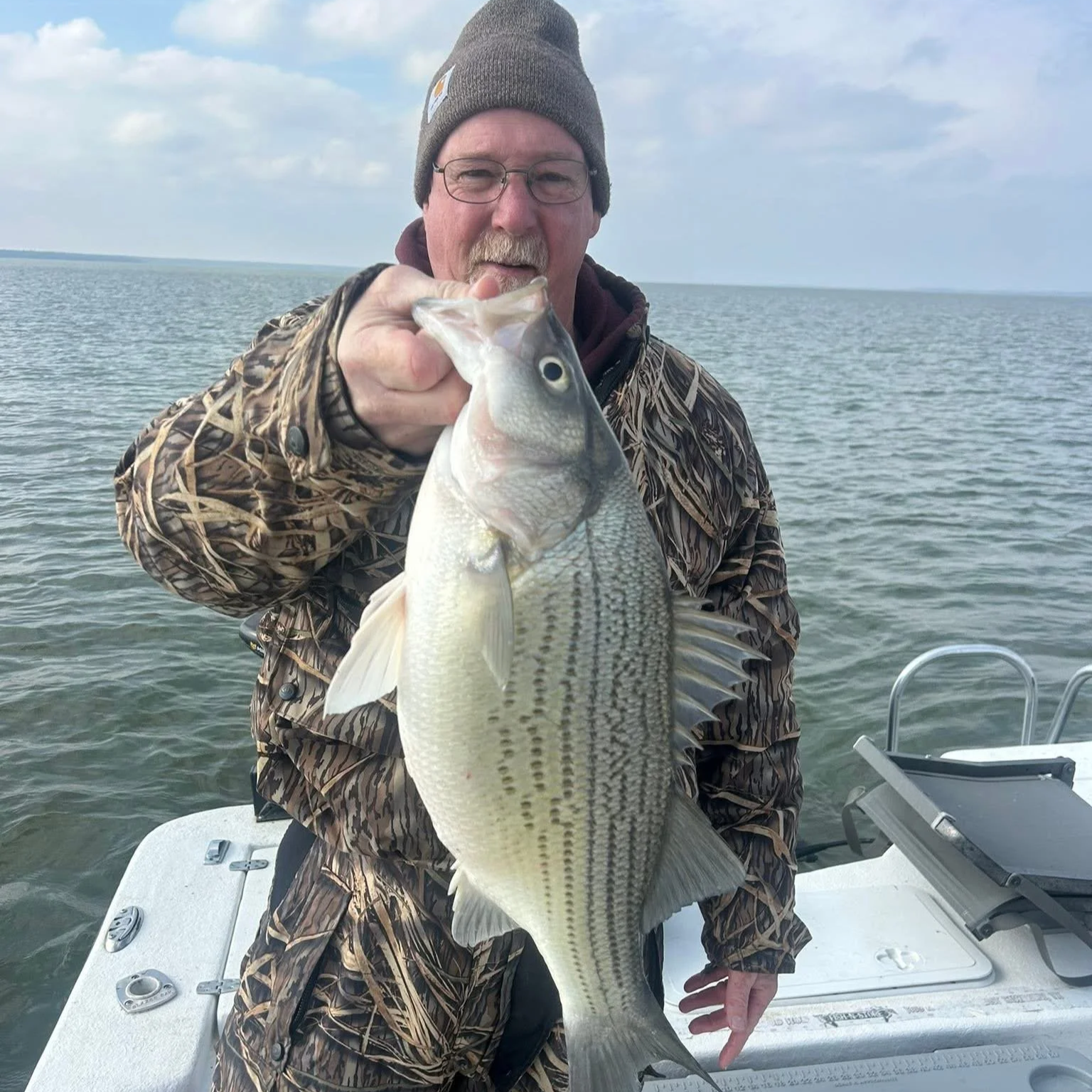 A man wearing a gray beanie, glasses, and camouflage jacket holding a large fish on a boat in open water on a cloudy day.