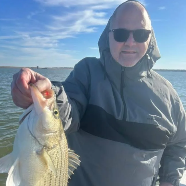 Bald man wearing sunglasses and a rain jacket holding a fish on a boat in a body of water under a blue sky with some clouds.
