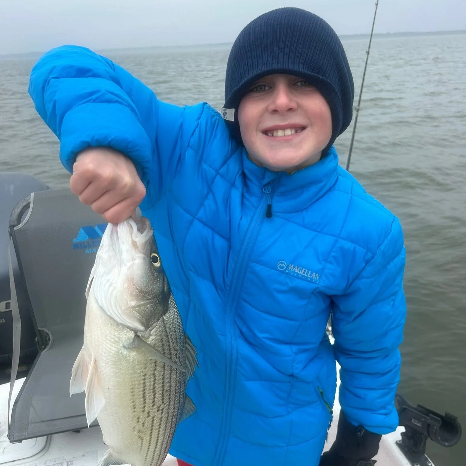 A person in a blue jacket and black beanie holding a fish on a boat in a body of water.