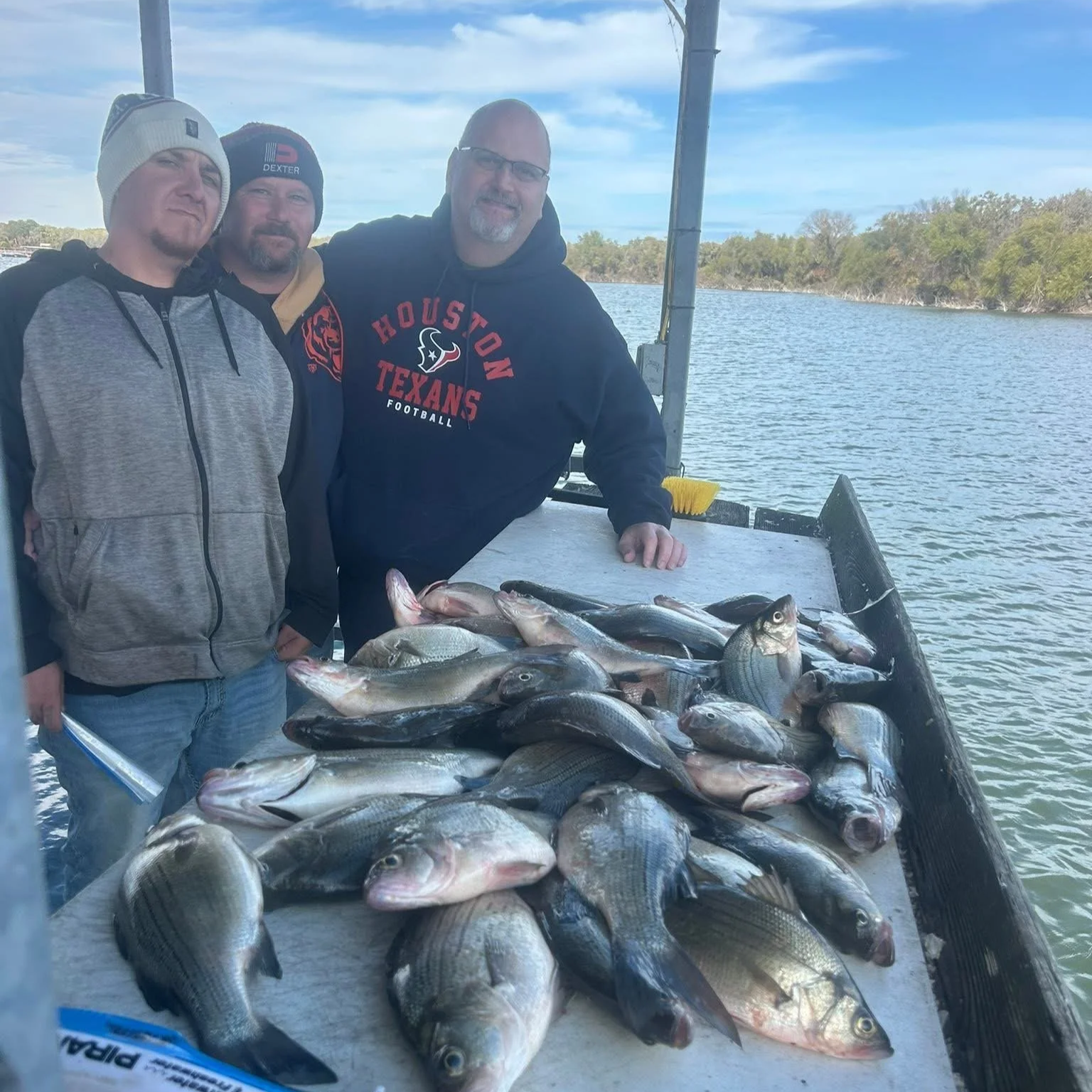 Three men standing on a boat with a large catch of fish on a table, with water and trees in the background.