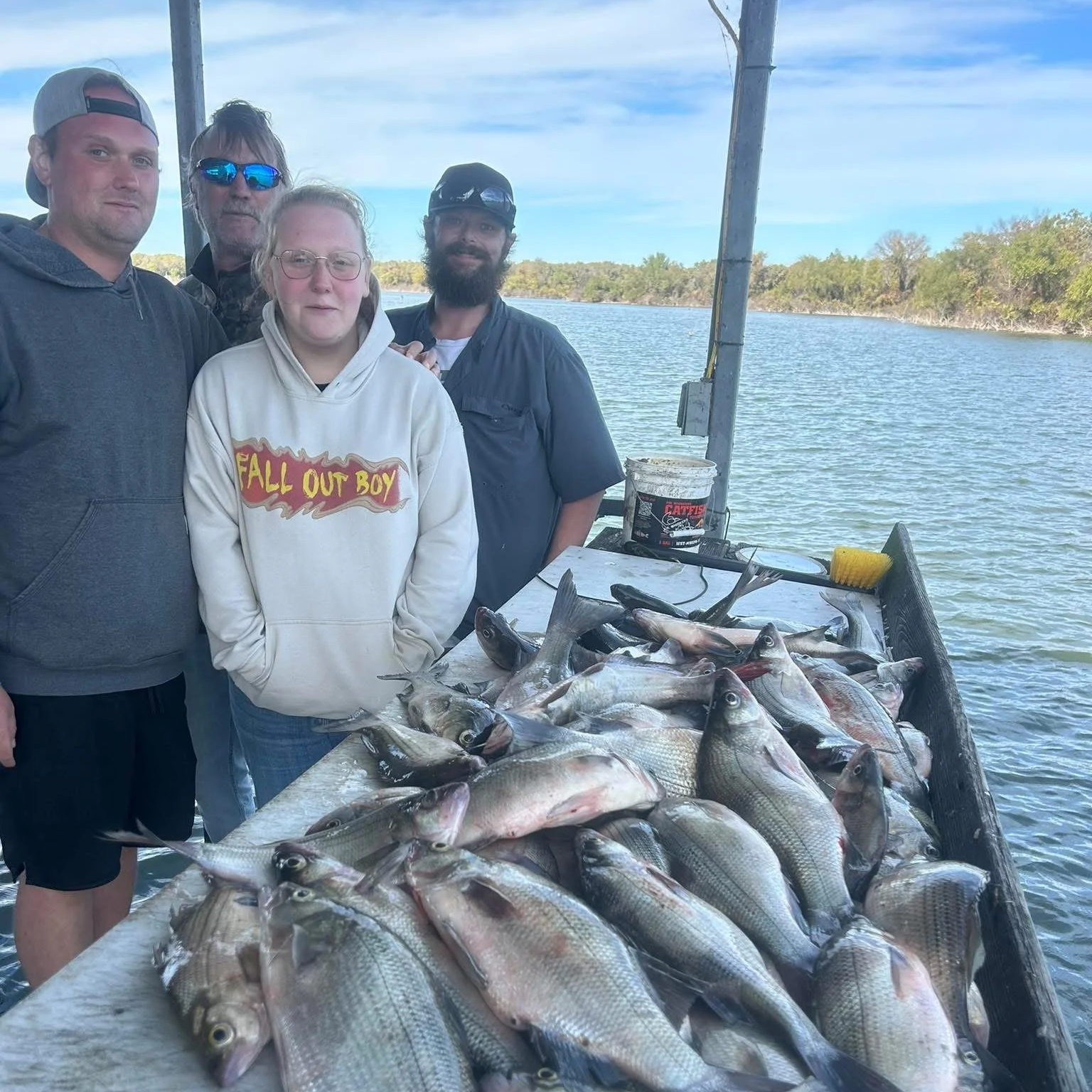 Four people standing behind a table filled with caught fish on a boat, with water and trees in the background during the daytime.