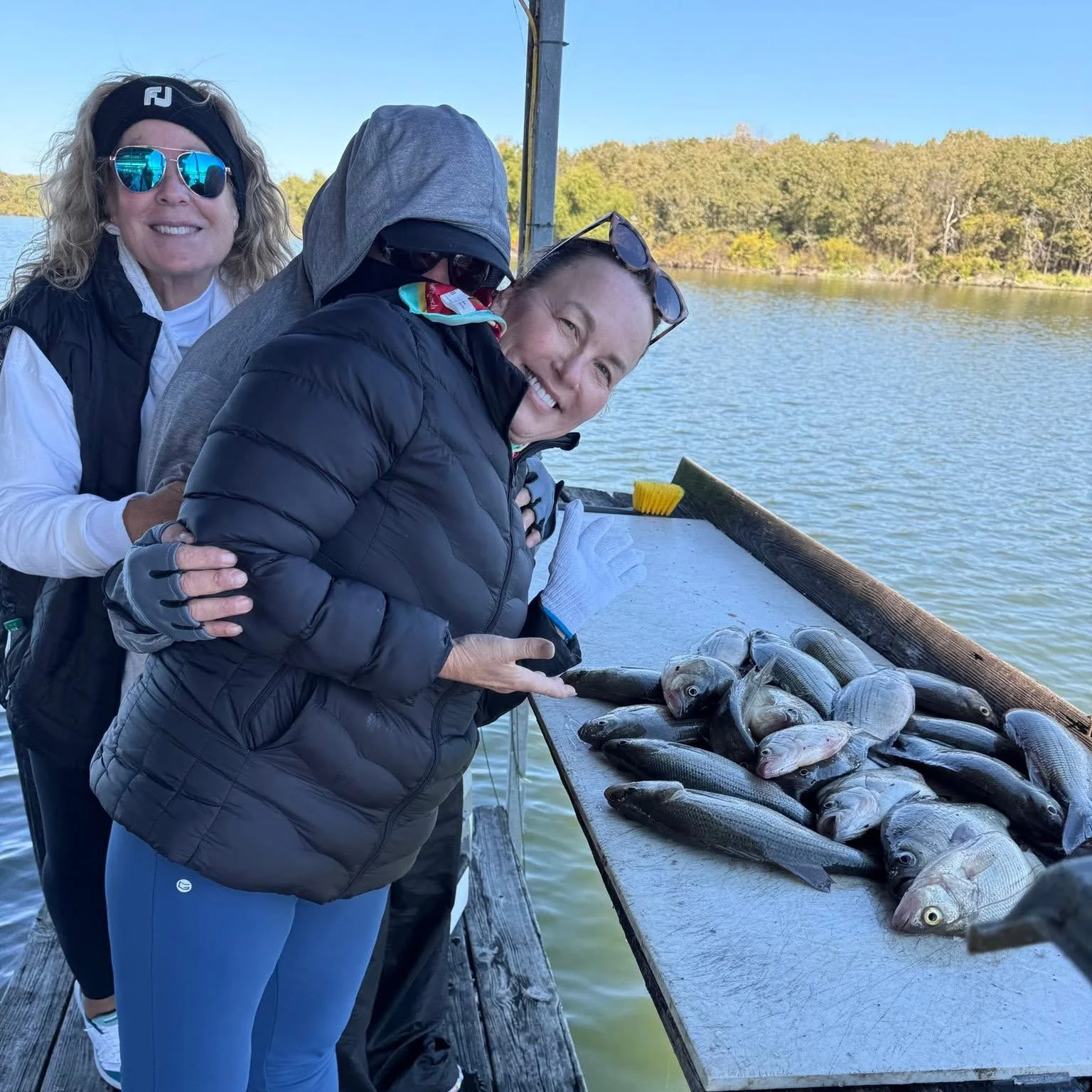 Two women smiling and holding a fish, standing next to a table with multiple fish on it by a body of water with trees in the background.
