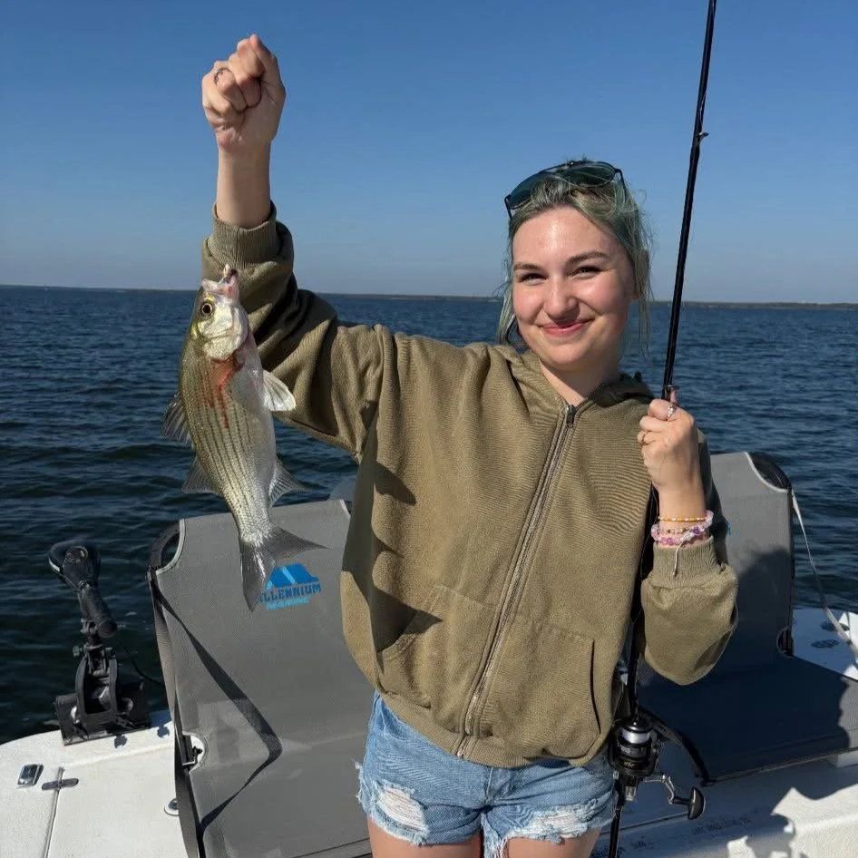 A young woman with blonde hair and a hoodie smiling while holding a fish she caught on a fishing line, on a boat in the water.