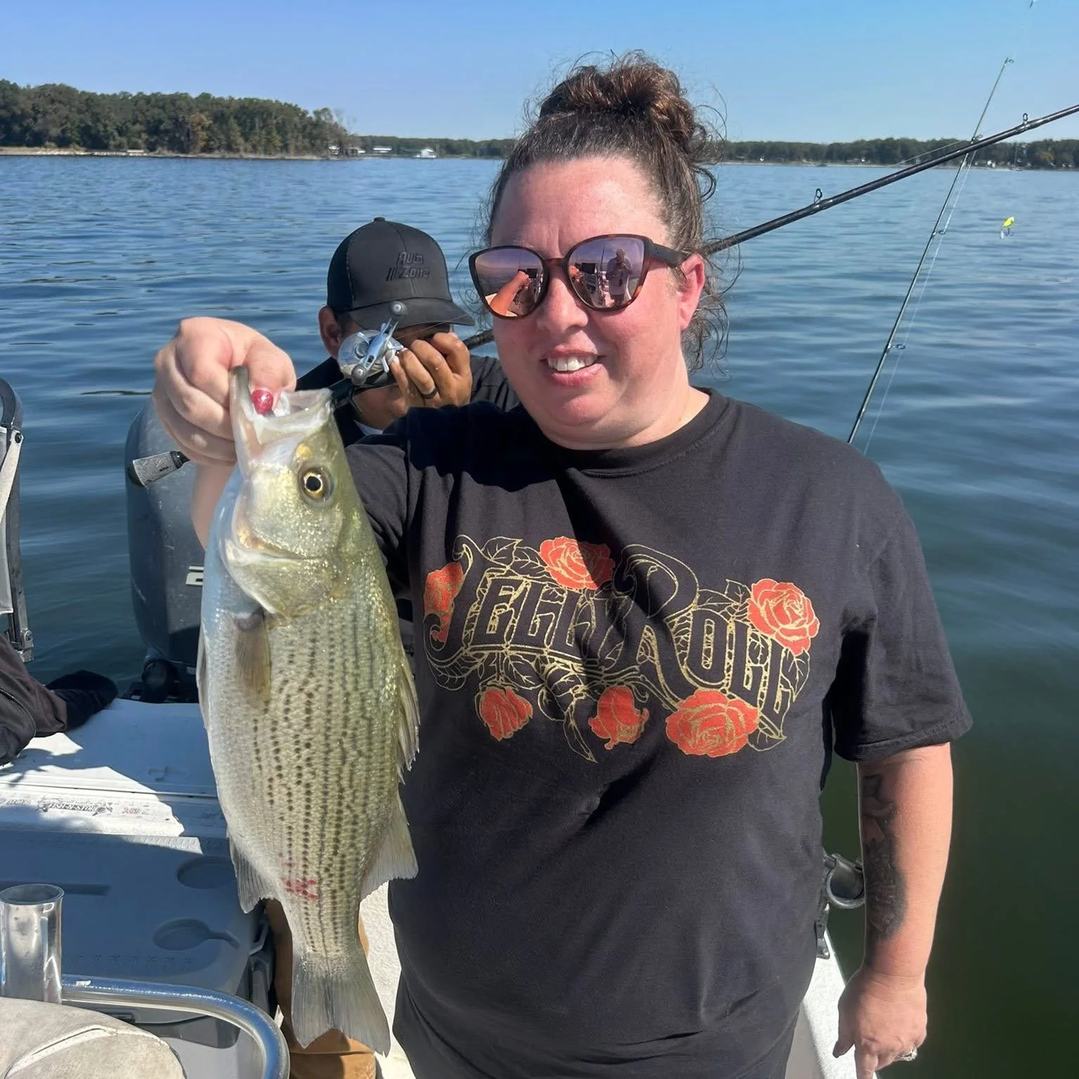 Woman smiling on a boat holding a large fish she caught, with person in the background and water and shoreline in the distance.