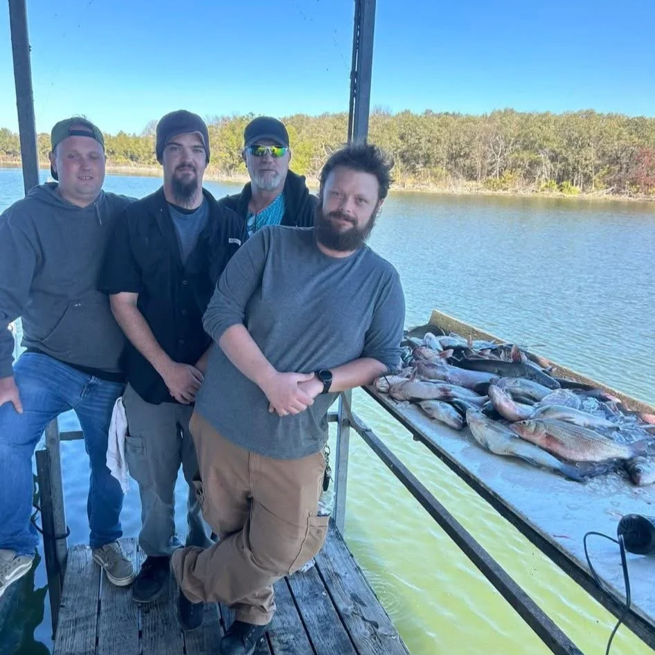 Four men standing on a dock near a body of water, with a table of freshly caught fish in front of them, and a wooded shoreline in the background.