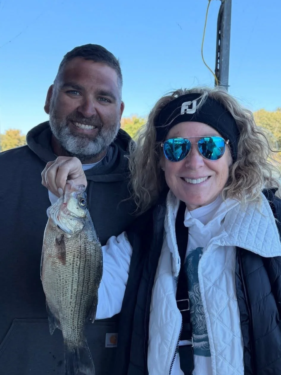 A man and a woman smiling outdoors, holding a large fish. The man has short dark hair and a beard, wearing a dark hoodie. The woman has curly blonde hair, wearing sunglasses, a headband, and a white and black vest. They are standing near a pole with 