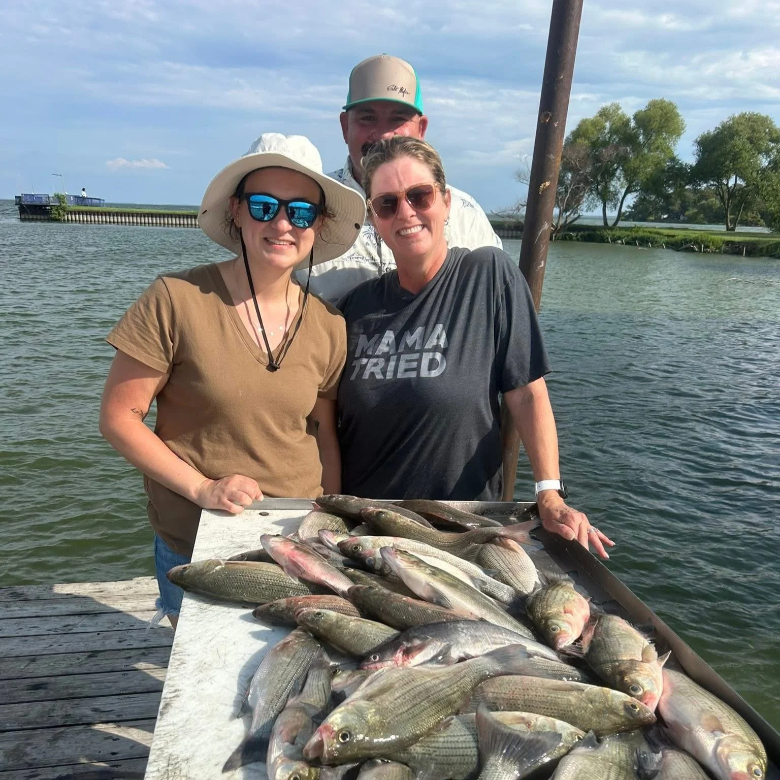 Three people standing behind a table filled with caught fish, on a dock near a body of water, with trees and a cloudy sky in the background.