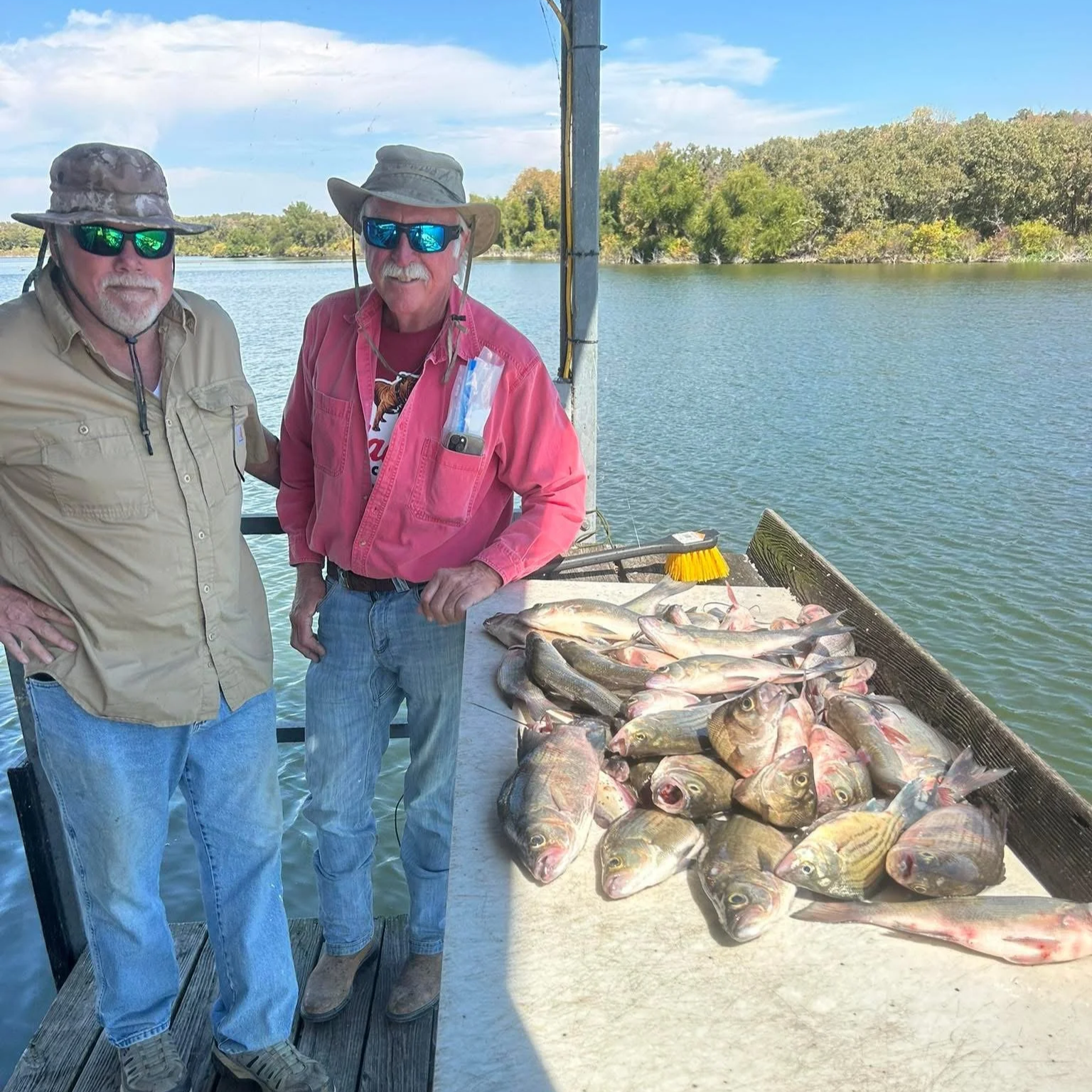 Two men standing on a dock with a catch of fish laid out on a table, near a body of water with trees in the background.
