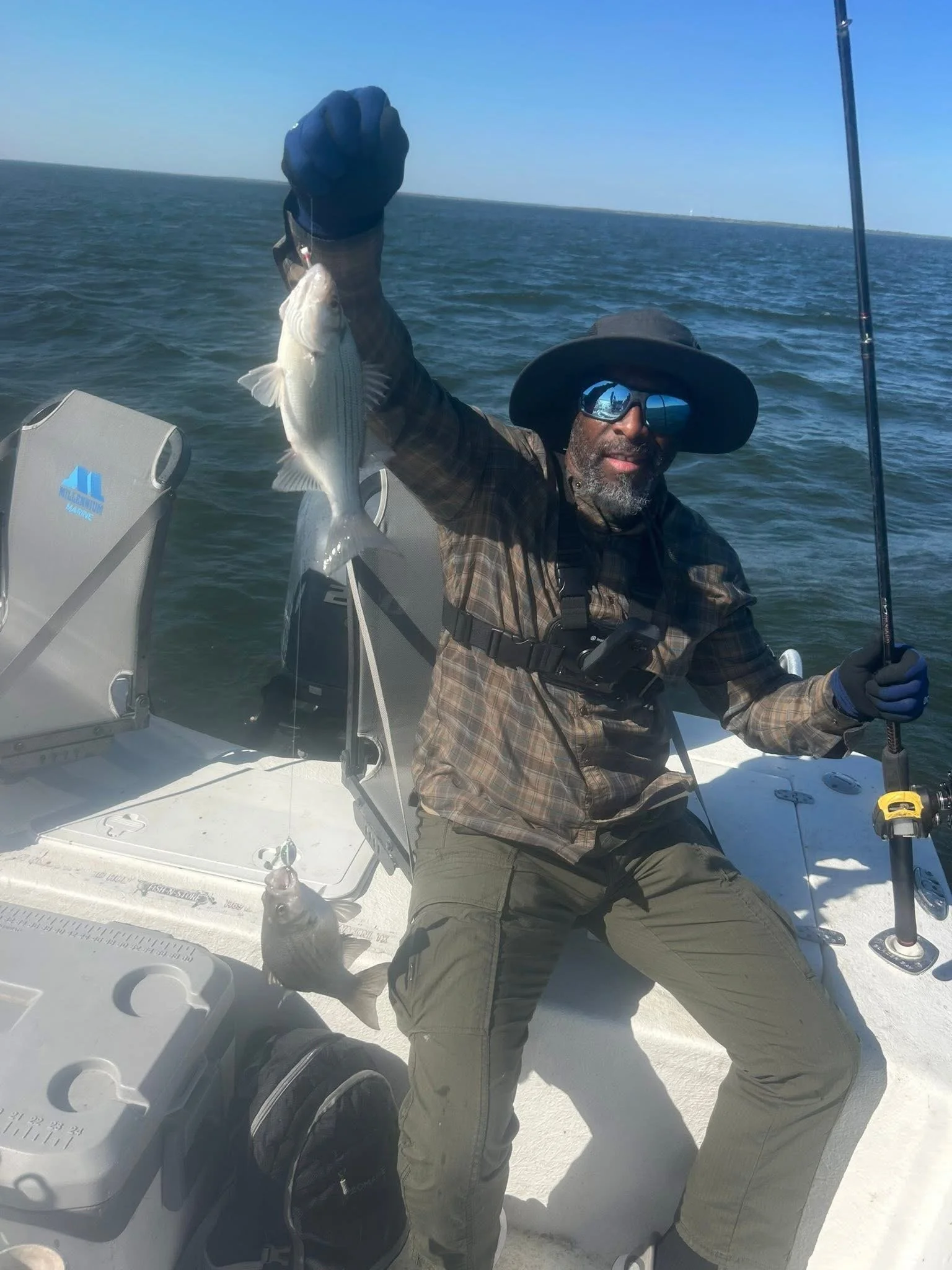 A man wearing sunglasses, a wide-brimmed hat, and outdoor clothing sits in a boat holding a fishing rod in one hand and a fish in the other, with open water and a distant shoreline in the background.
