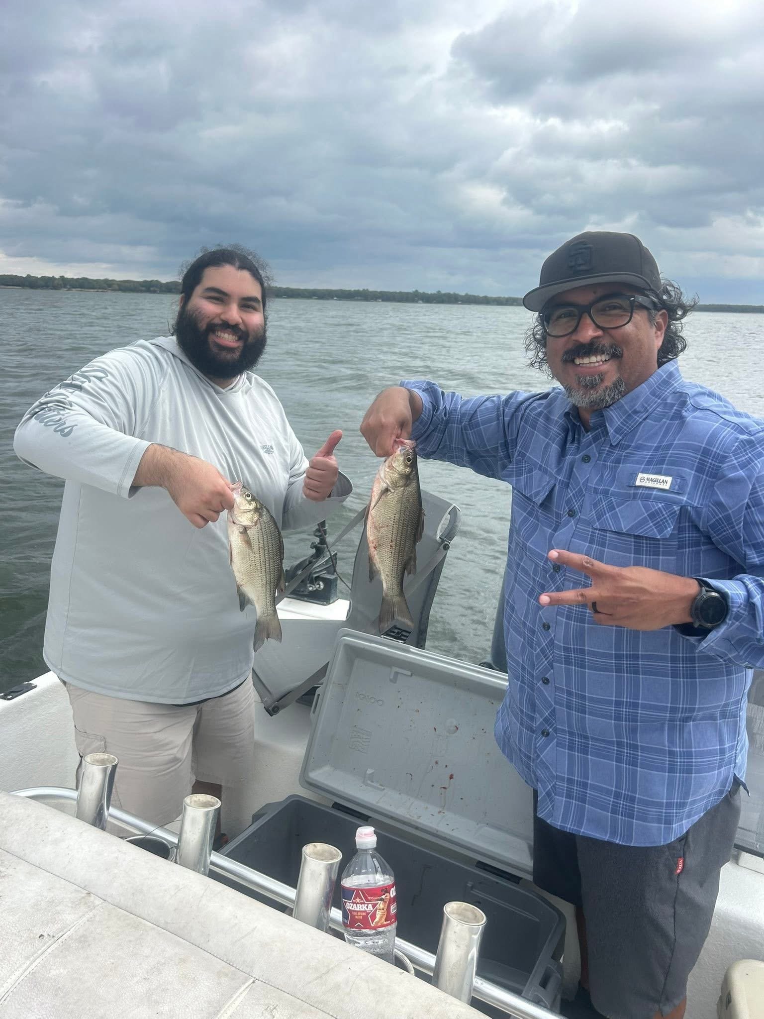 Two men on a boat holding fish and smiling, with a body of water and cloudy sky in the background.