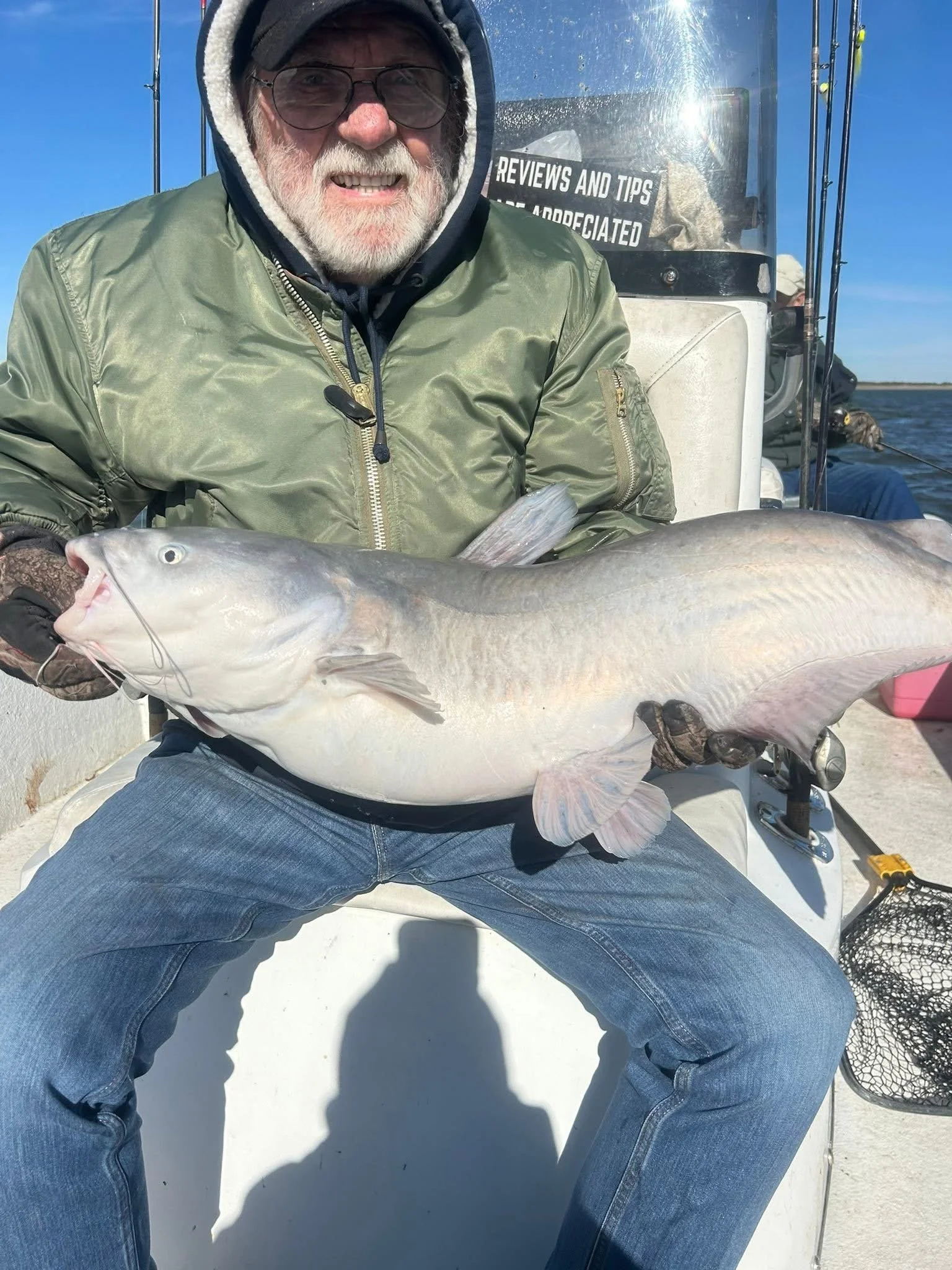 An older man with glasses and a beard, wearing a hoodie and jacket, sitting on a boat and holding a large fish in his lap. There is a person in the background fishing, and a sign on the boat that reads 'REVIEWS AND TIPS'. The sky is clear and blue.
