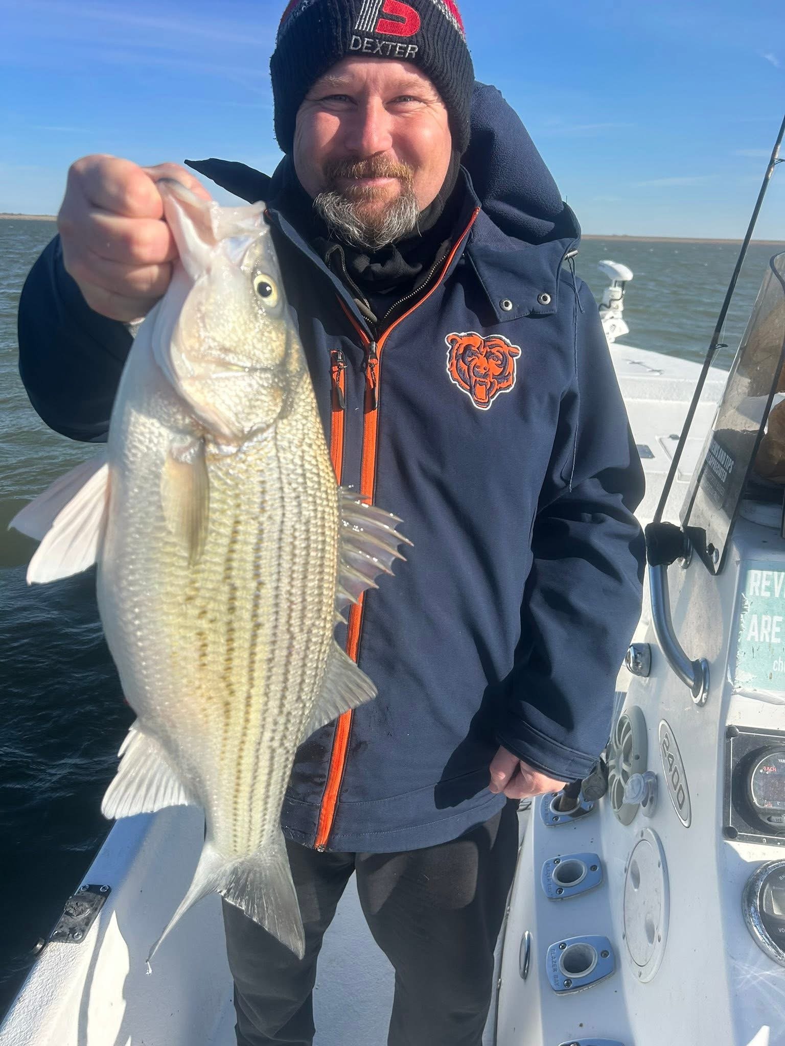A man with a beard and mustache wearing a black beanie and dark jacket, holding a fish with a yellowish body and a dark eye, on a boat in a body of water under a blue sky.