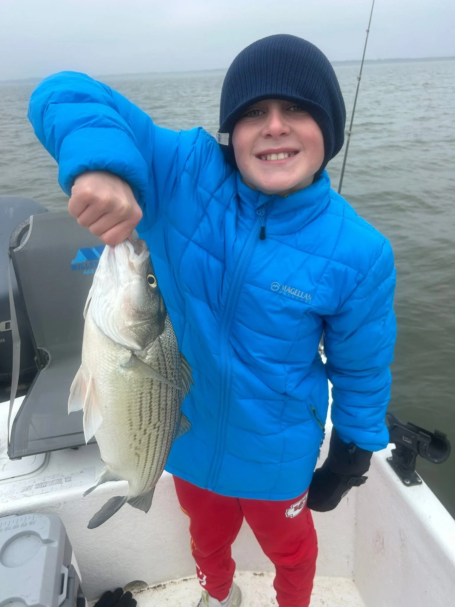 A person dressed in a blue jacket and red pants holding a large fish on a boat. The person is smiling and wearing a dark blue beanie. The water and cloudy sky are visible in the background.