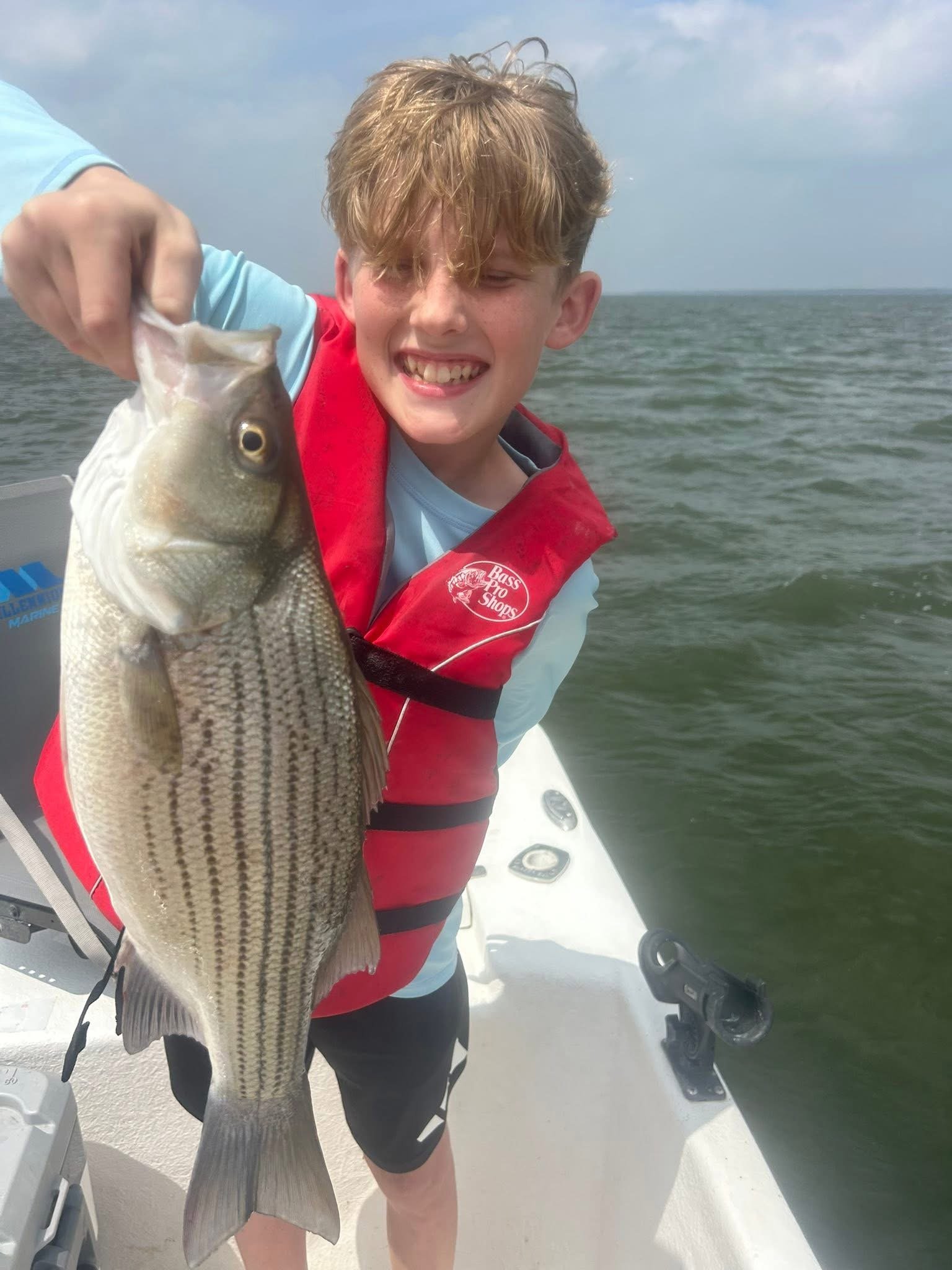 A boy holding a large fish on a boat with water and cloudy sky in the background.