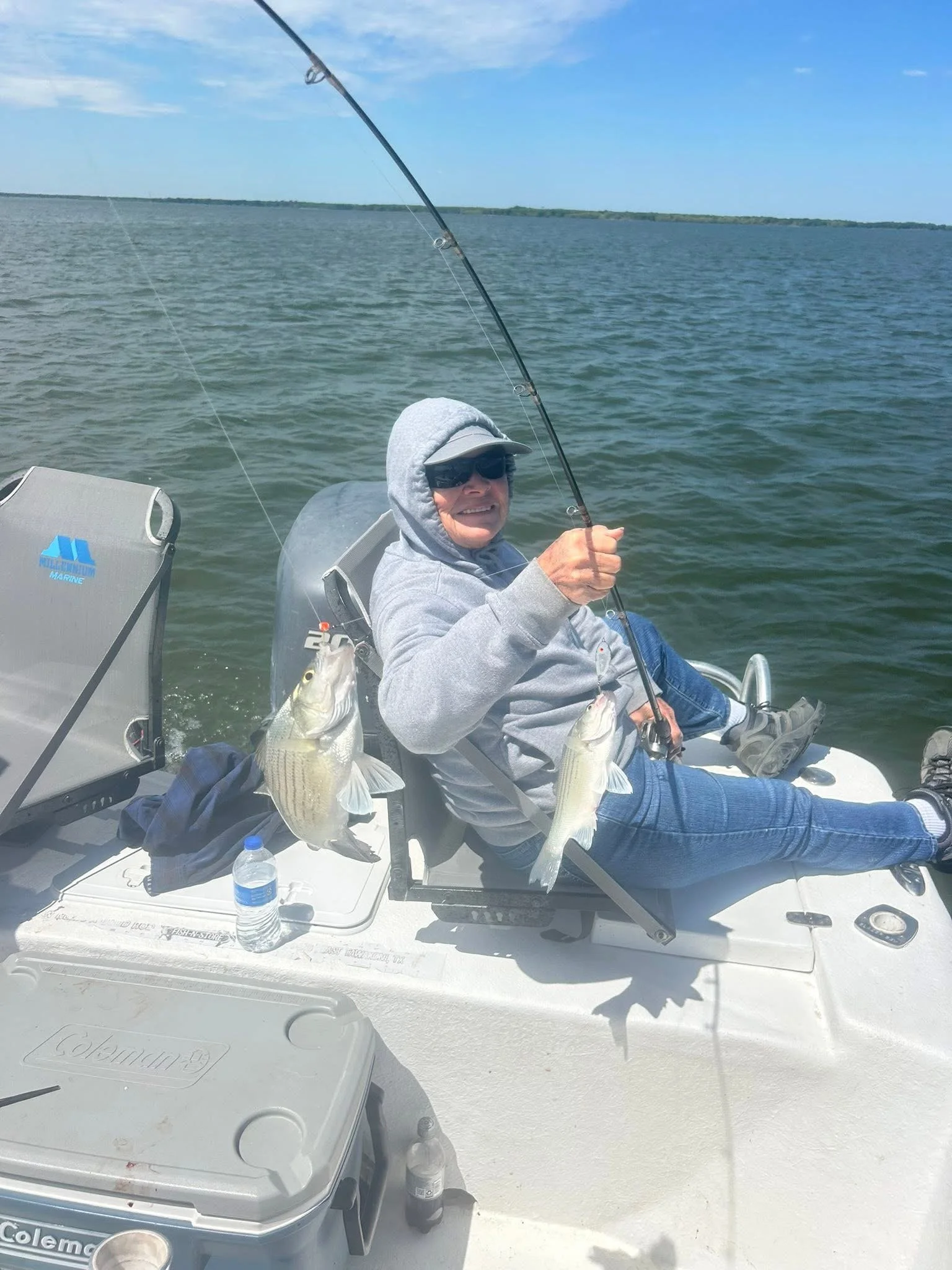 An elderly woman sitting on a boat holding two fish she caught while fishing on a lake under a blue sky.