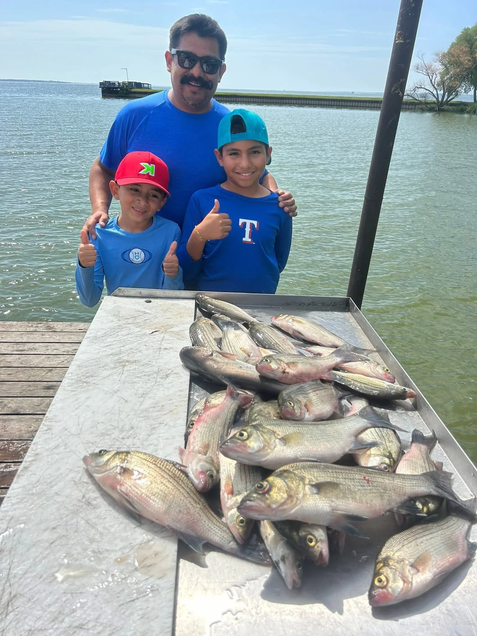 Three people, an adult man and two boys, standing behind a table of freshly caught fish near a body of water, giving thumbs-up signs.