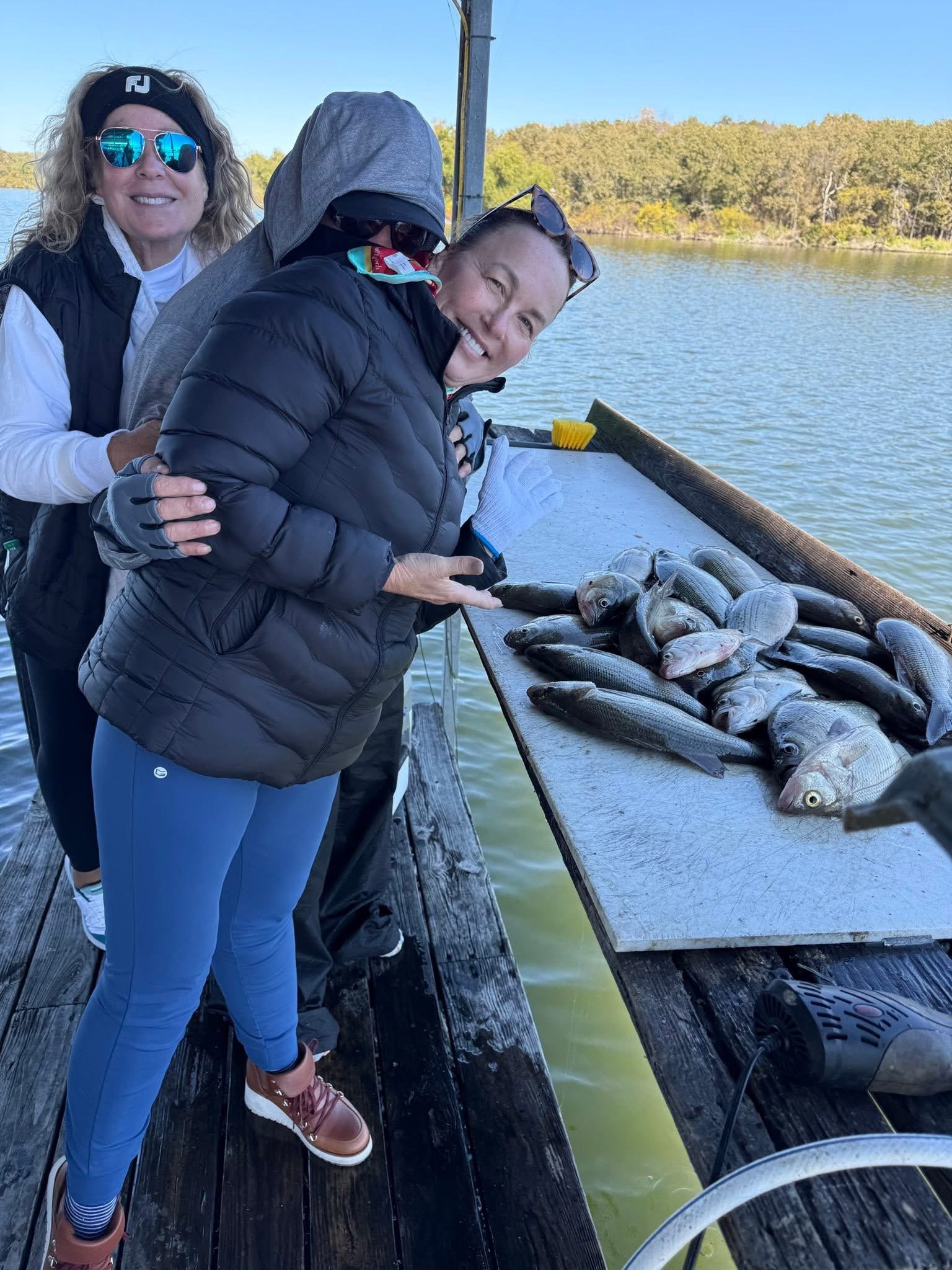 Three women on a dock by the water, smiling, with a table of fish, during daytime. One woman is wearing sunglasses, a black vest, and blue leggings. The second woman, partially hidden, wears a hoodie. The third woman, in front, wears a black puffer jacket and sunglasses on her head, holding a fish.