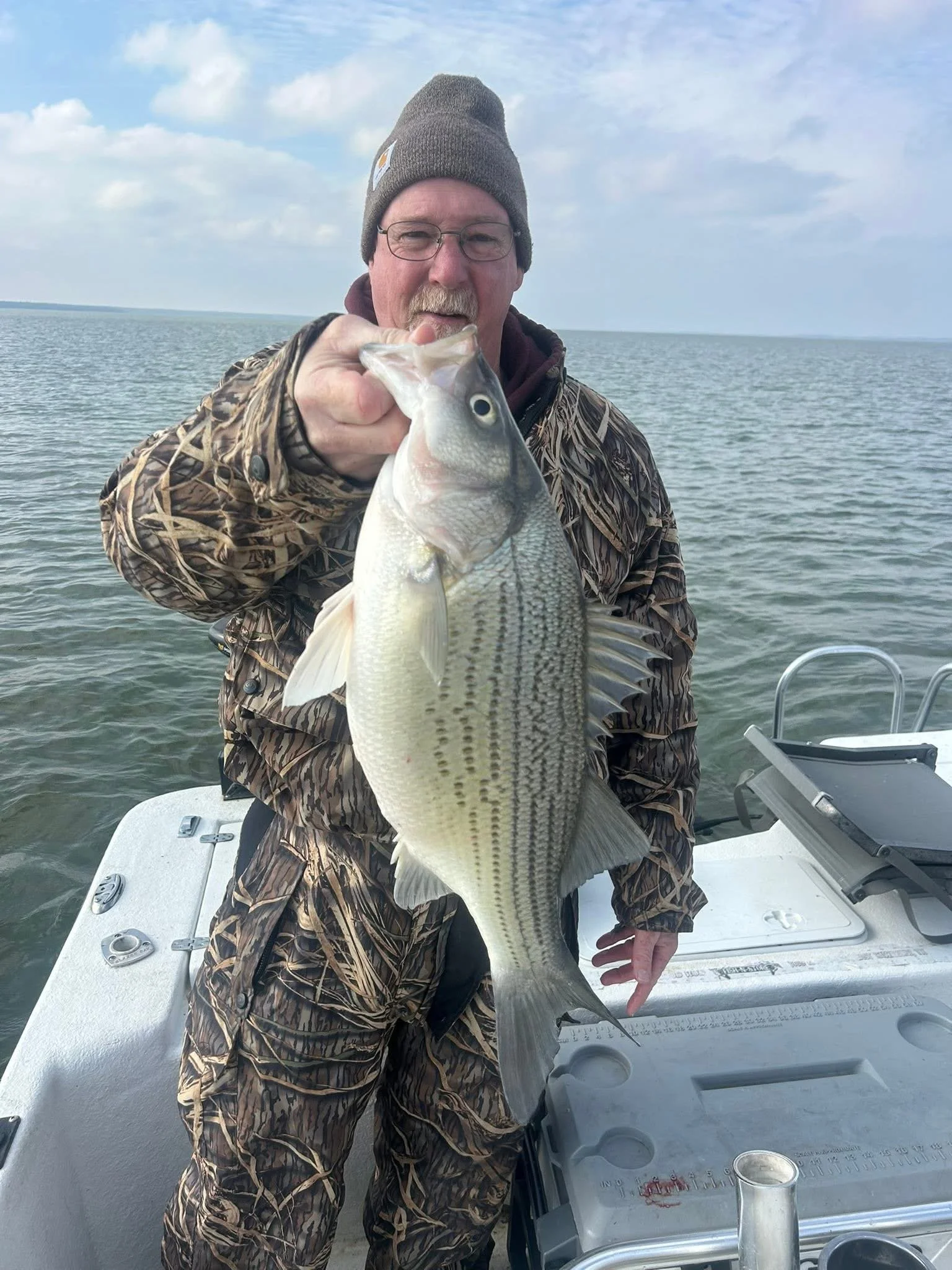 A man with glasses, wearing a gray beanie and camouflage jacket, is holding a large fish on a boat in the water, with the ocean and sky in the background.