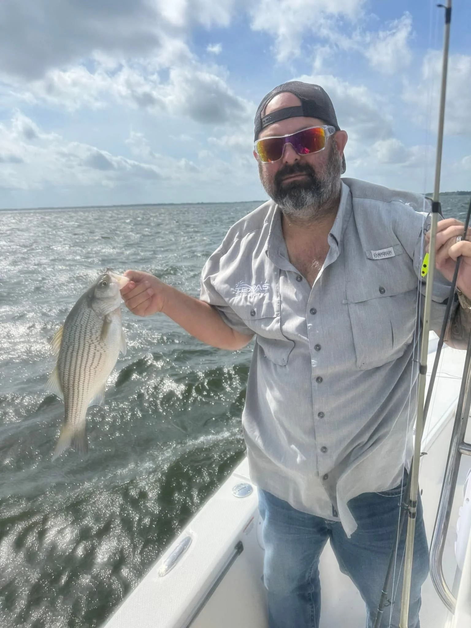 A man with a beard, wearing sunglasses, a black cap, and a gray fishing shirt, holding a fish on a boat with water and sky in the background.