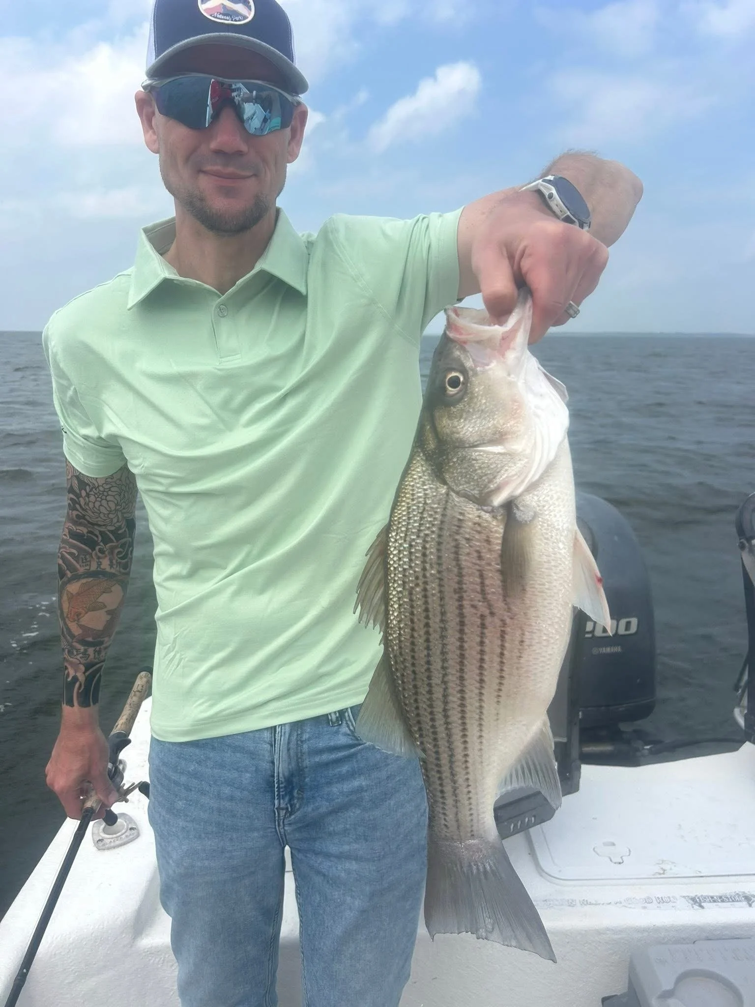 A man in a green shirt and jeans holding a large fish on a boat with water and sky in the background.