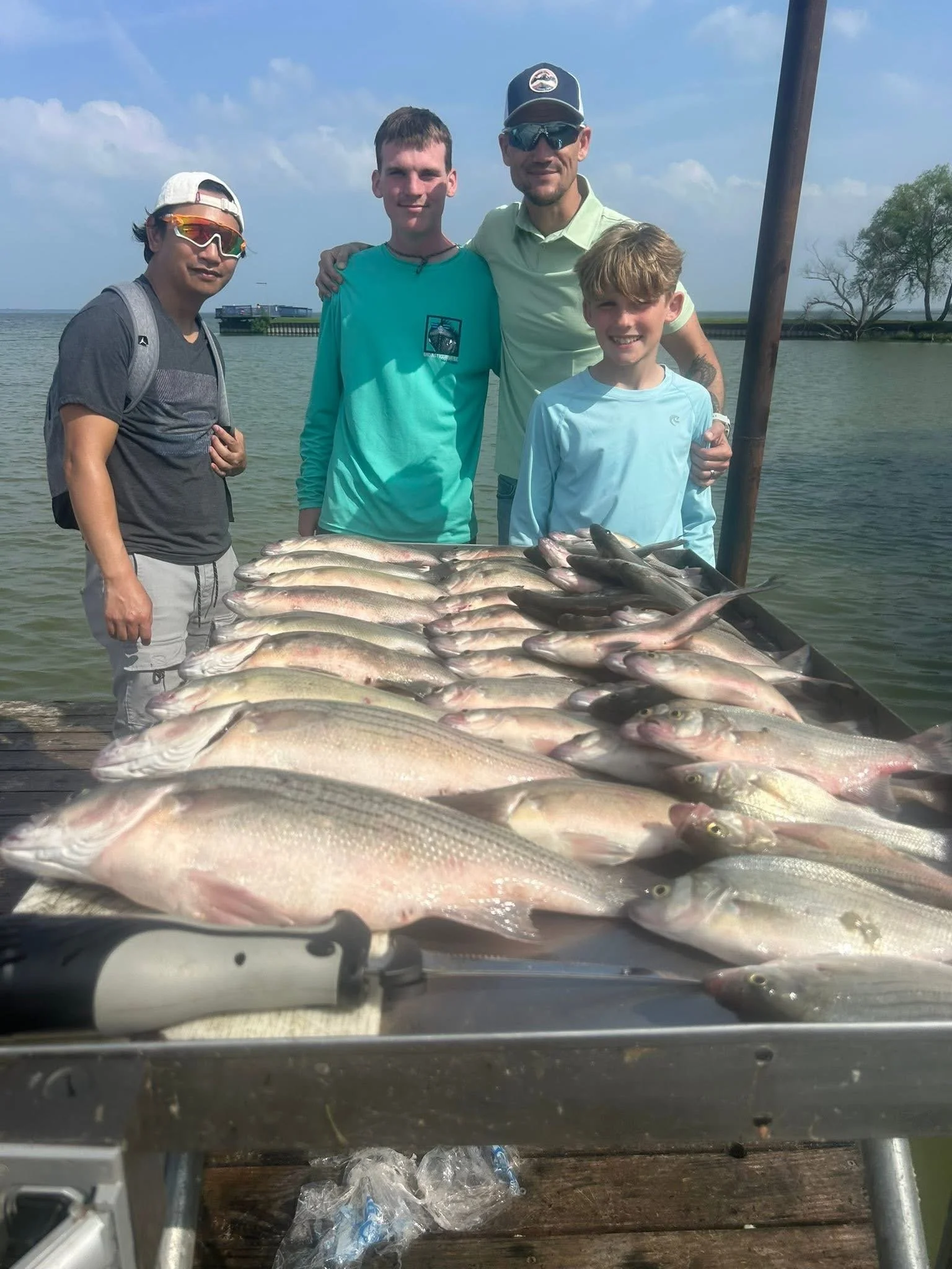 Four people are standing behind a large display of fish. The fish are arranged on a surface, possibly a boat or dock. The group is near a body of water with a pier, trees, and a cloudy sky in the background.