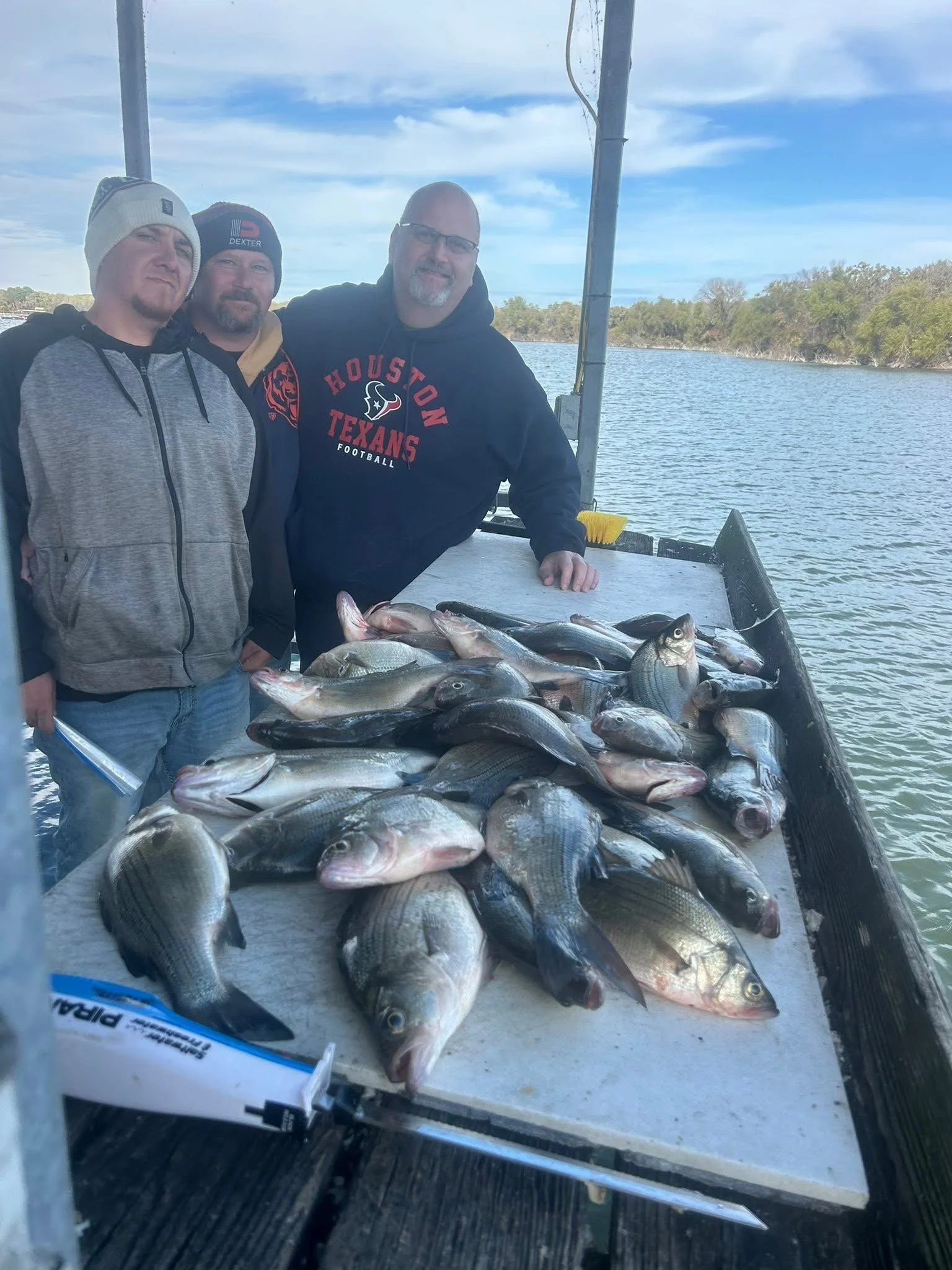 Three men on a boat displaying a large catch of fish, with water and treeline in the background.