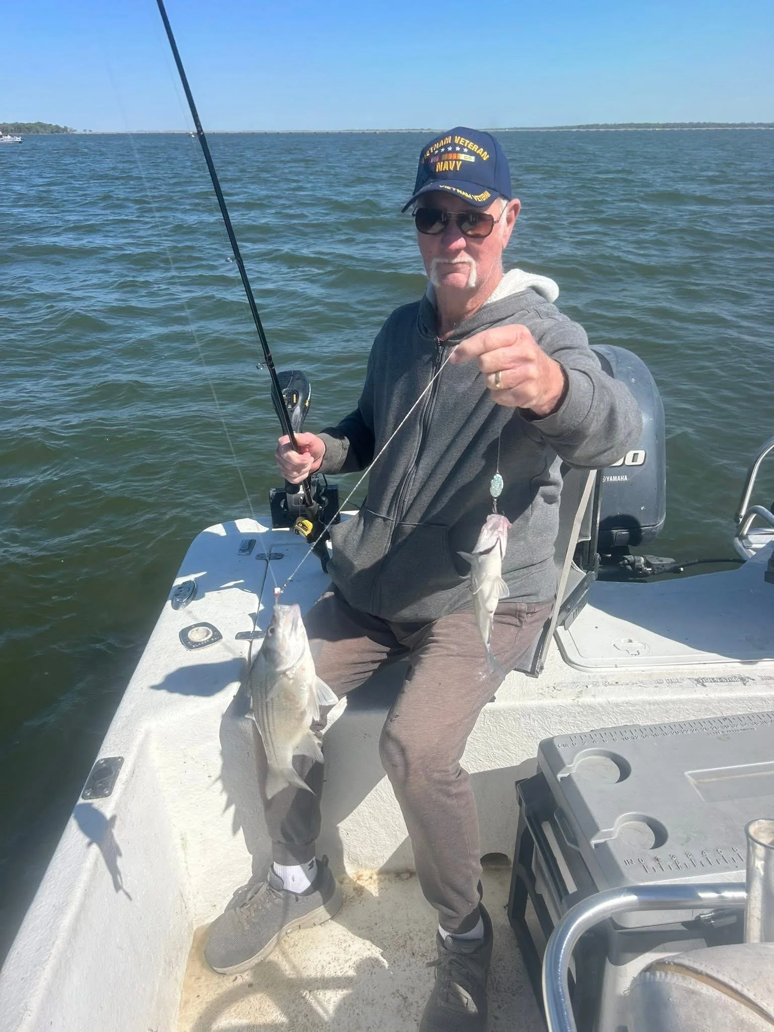 An older man wearing sunglasses, a navy veteran hat, and a gray hoodie sitting in a boat on the water, holding a fishing rod with two fish caught.