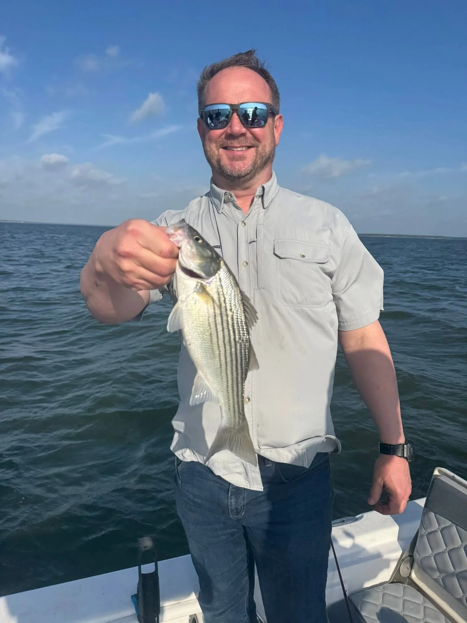 A man with short brown hair, beard, and sunglasses holding a fish he caught while fishing on a boat in the ocean under a blue sky.