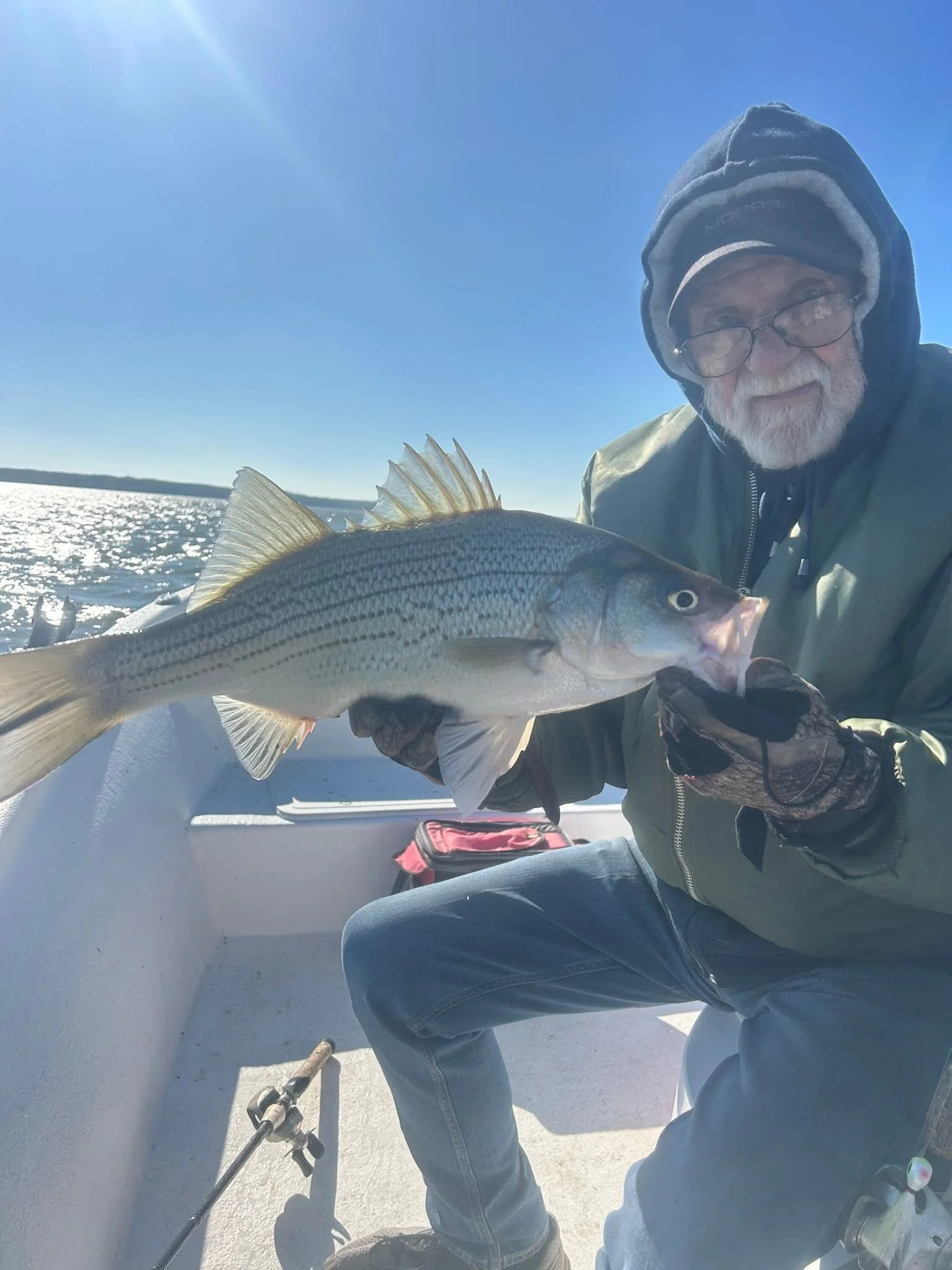 Older man with a gray beard, glasses, and a hooded jacket holds a large fish on icy ground with a body of water and blue sky in the background.
