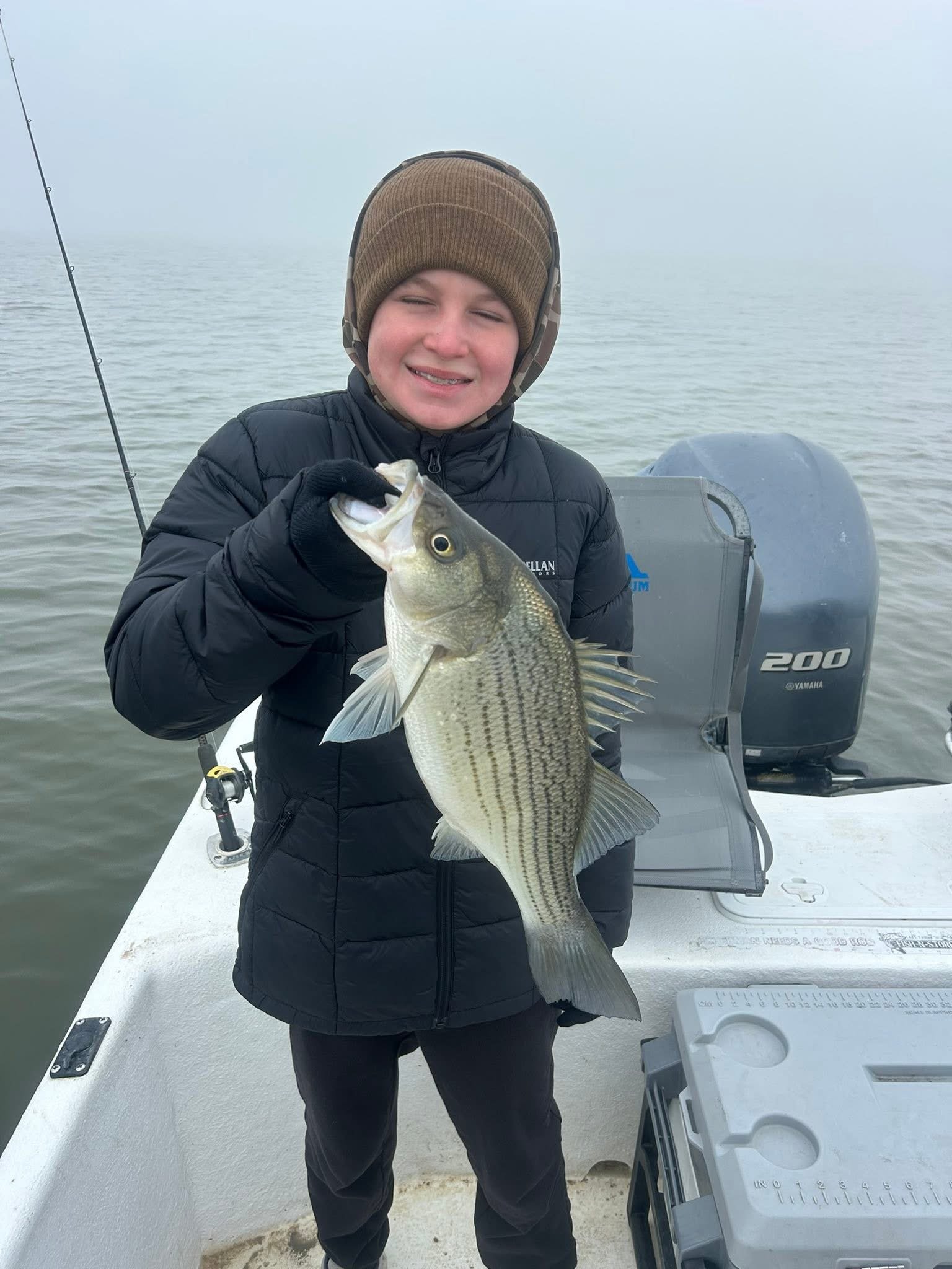 A young boy on a boat holding a large fish, wearing a brown beanie, black jacket, and black gloves, with water and fog in the background.