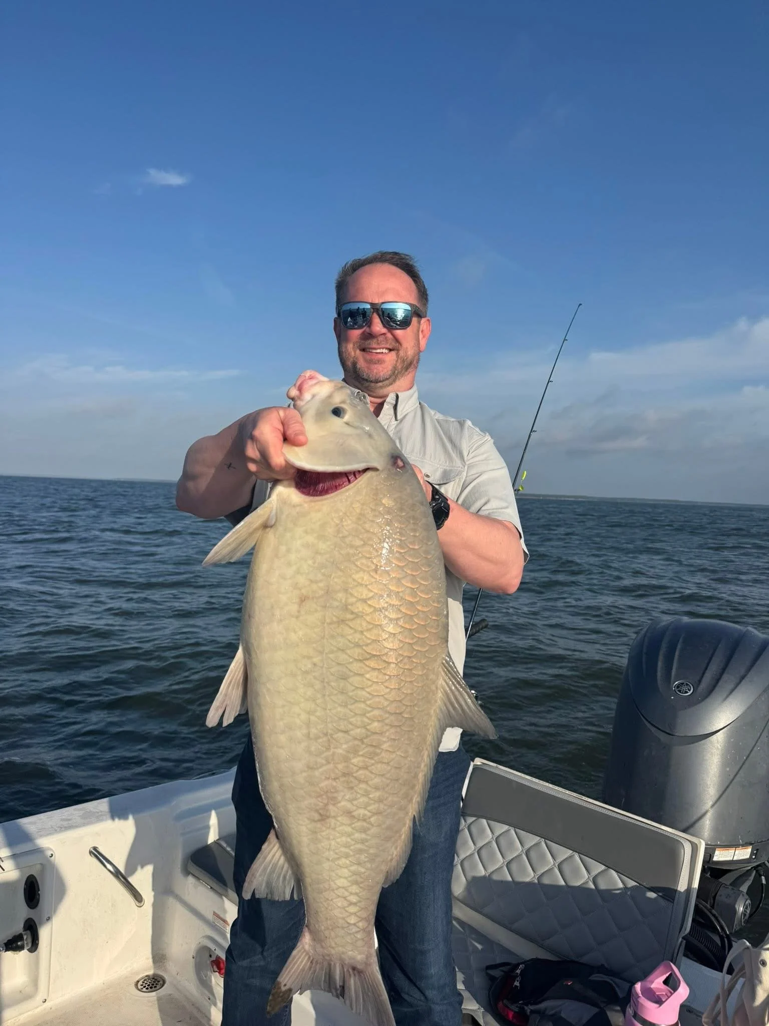 A man wearing sunglasses and a light-colored shirt holding up a large fish on a boat in the water. The background shows a blue sky with some clouds and the open water.