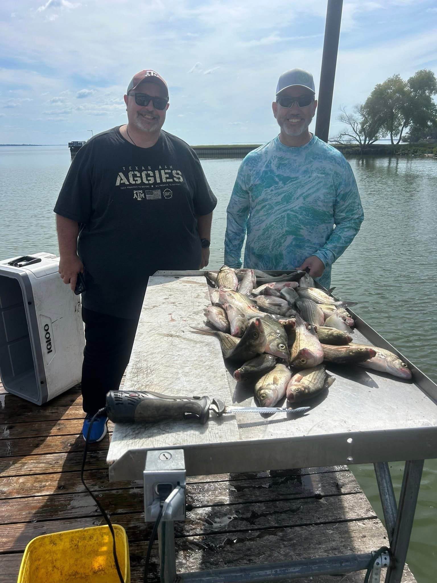 Two smiling men with sunglasses and caps standing behind a large table of freshly caught fish on a dock by a body of water, with trees and a pier in the background under a partly cloudy sky.